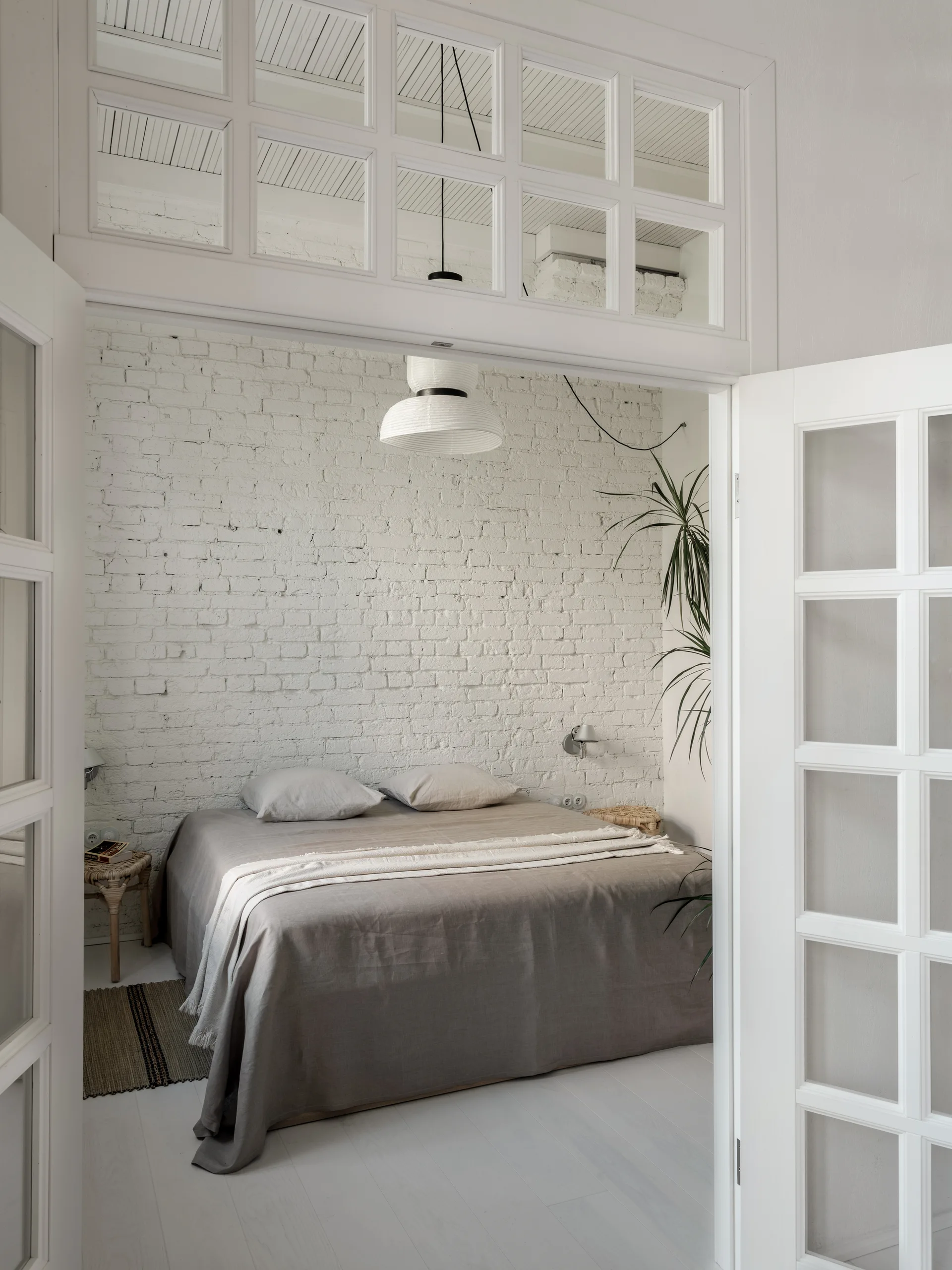 A bedroom with grey-beige linen bedding and a white brick wall is viewed through an open white French door with a matching transom window.