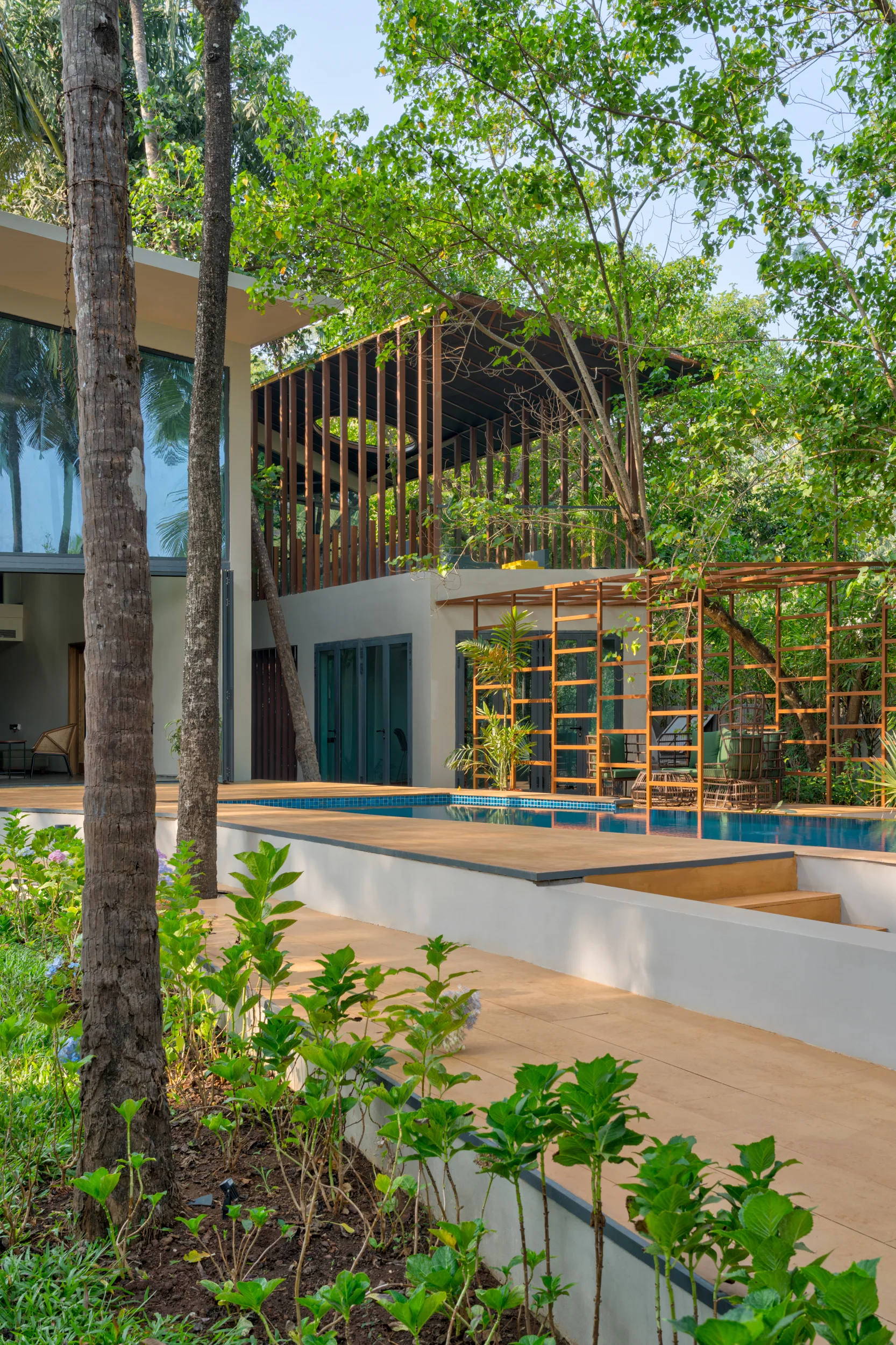 A modern outdoor lap pool and tan stone deck are nestled among mature green trees and a geometric wooden pergola structure.