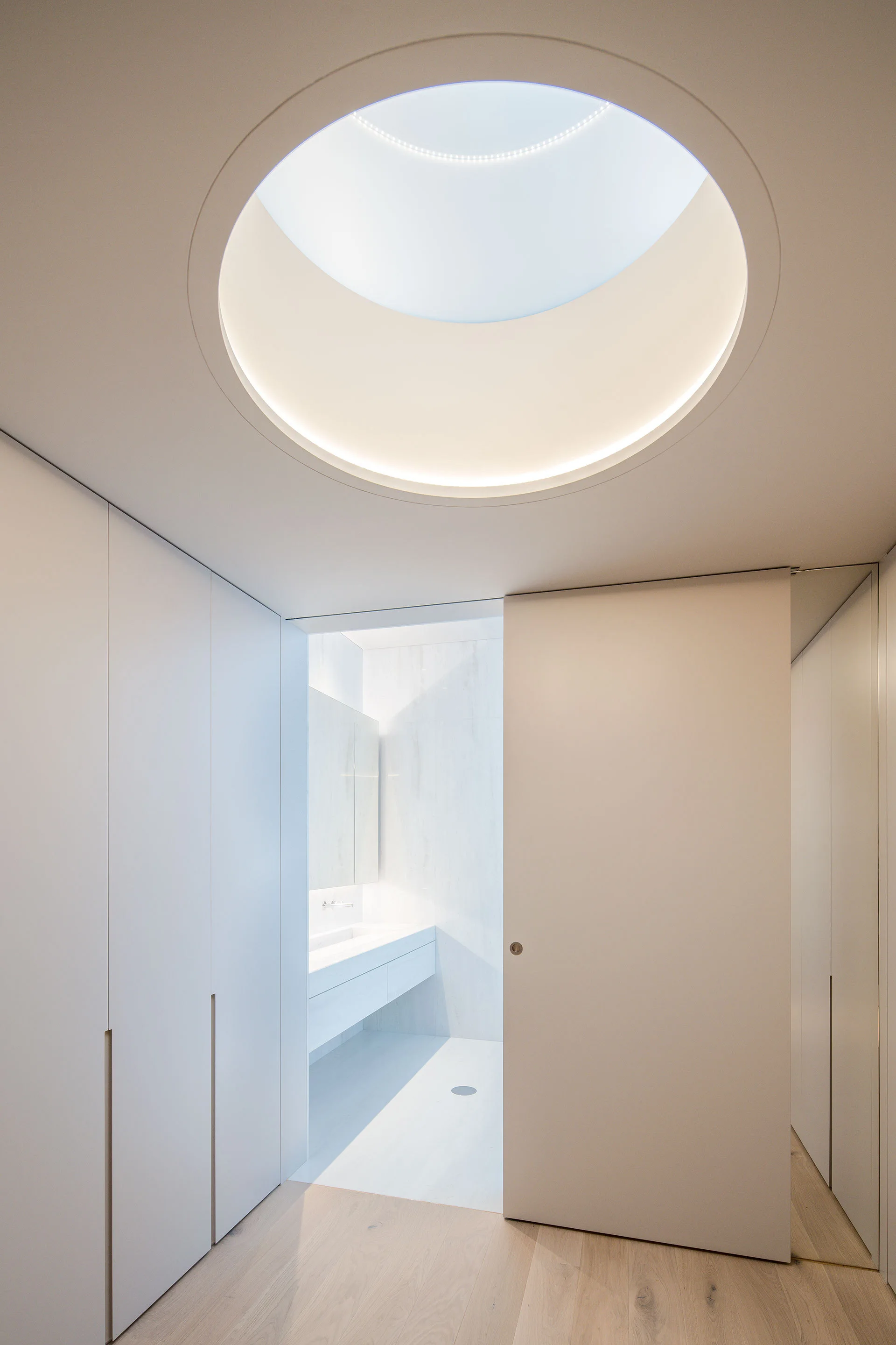 A minimalist white hallway features a large circular skylight overhead and a sliding door opening into a modern bathroom with a floating vanity.