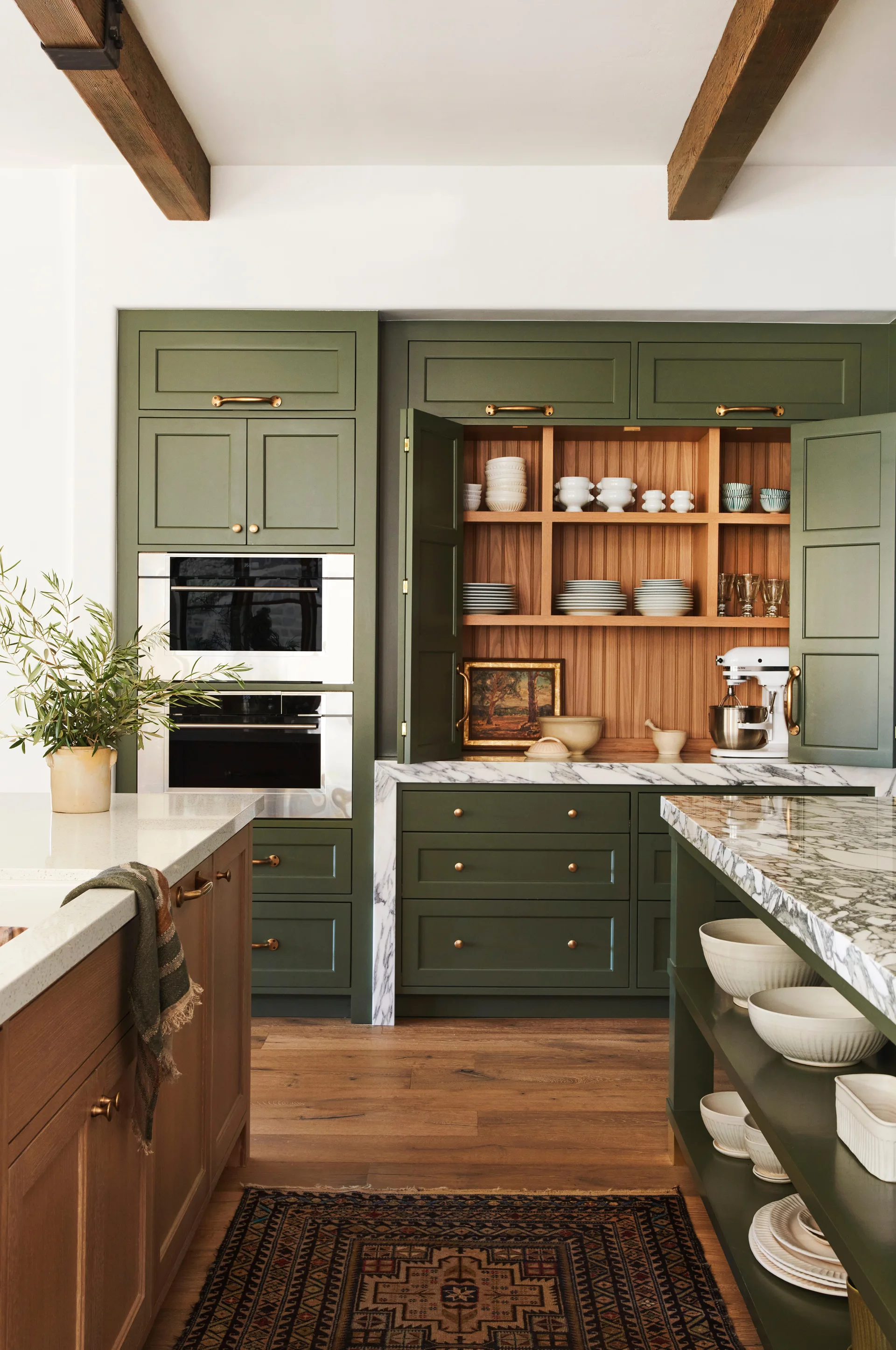 Olive green kitchen cabinets feature a built-in wood-lined pantry, marble countertops, and dark wood ceiling beams above a patterned rug.