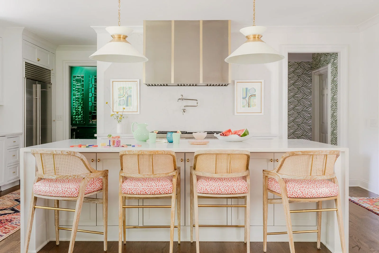 Four cane-back counter stools with patterned pink cushions sit at a white kitchen island beneath two pendant lights and a brass-trimmed range hood.