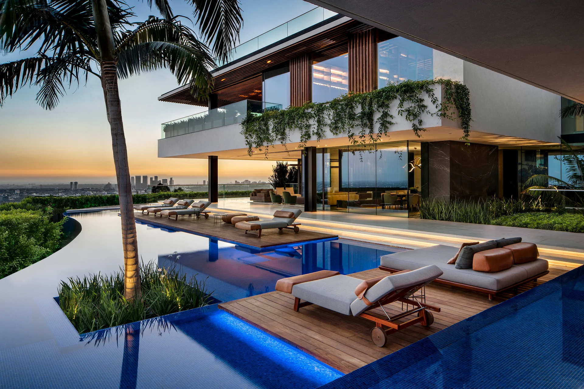 Modern multi-level home with an infinity pool featuring inset wood decks, sun loungers, and trailing balcony greenery at sunset.