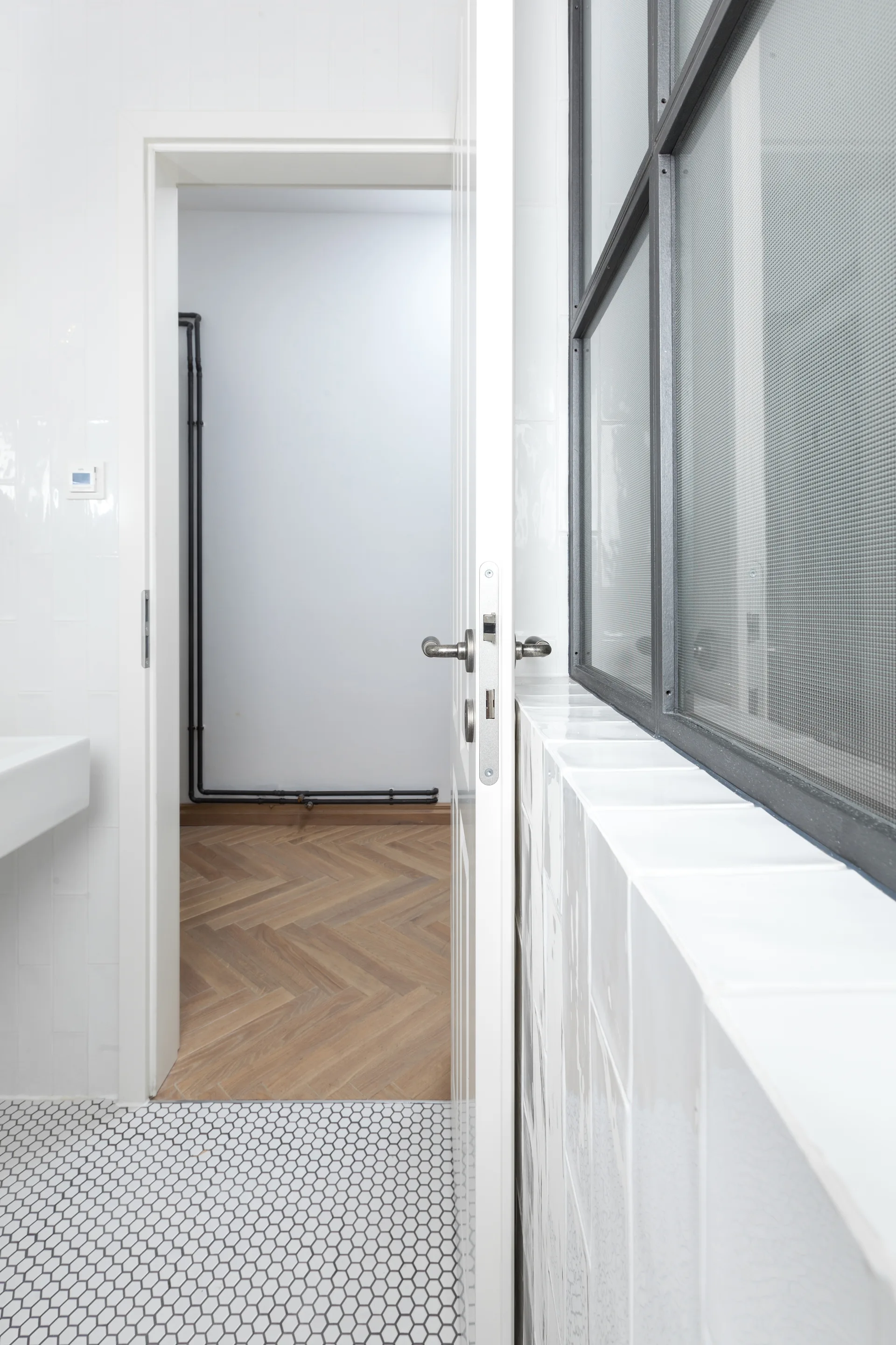 An open white door shows a transition from a white tiled bathroom floor to warm wood herringbone parquet flooring in the adjacent room.