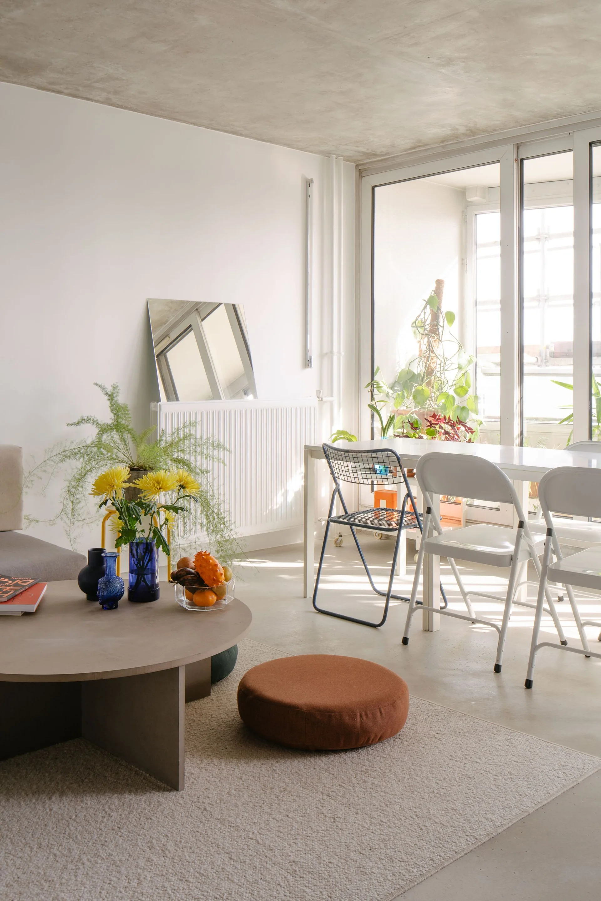 A bright open-plan living and dining area with an exposed concrete ceiling, a low round coffee table, and a white dining table with folding chairs.