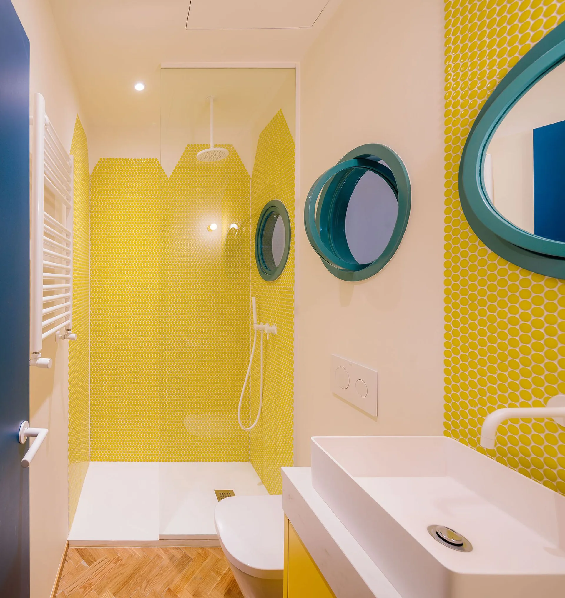 A modern bathroom features bright yellow penny tile walls, two turquoise circular mirrors, a white rectangular washbasin, and herringbone wood floors.