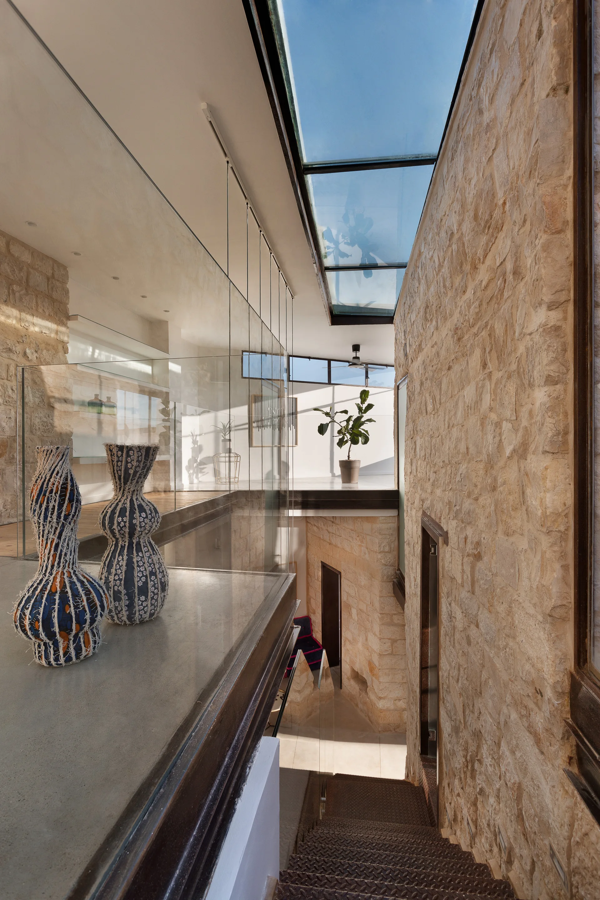 An interior view looking down a metal staircase flanked by a textured stone wall, glass railings, and an overhead glass skylight.