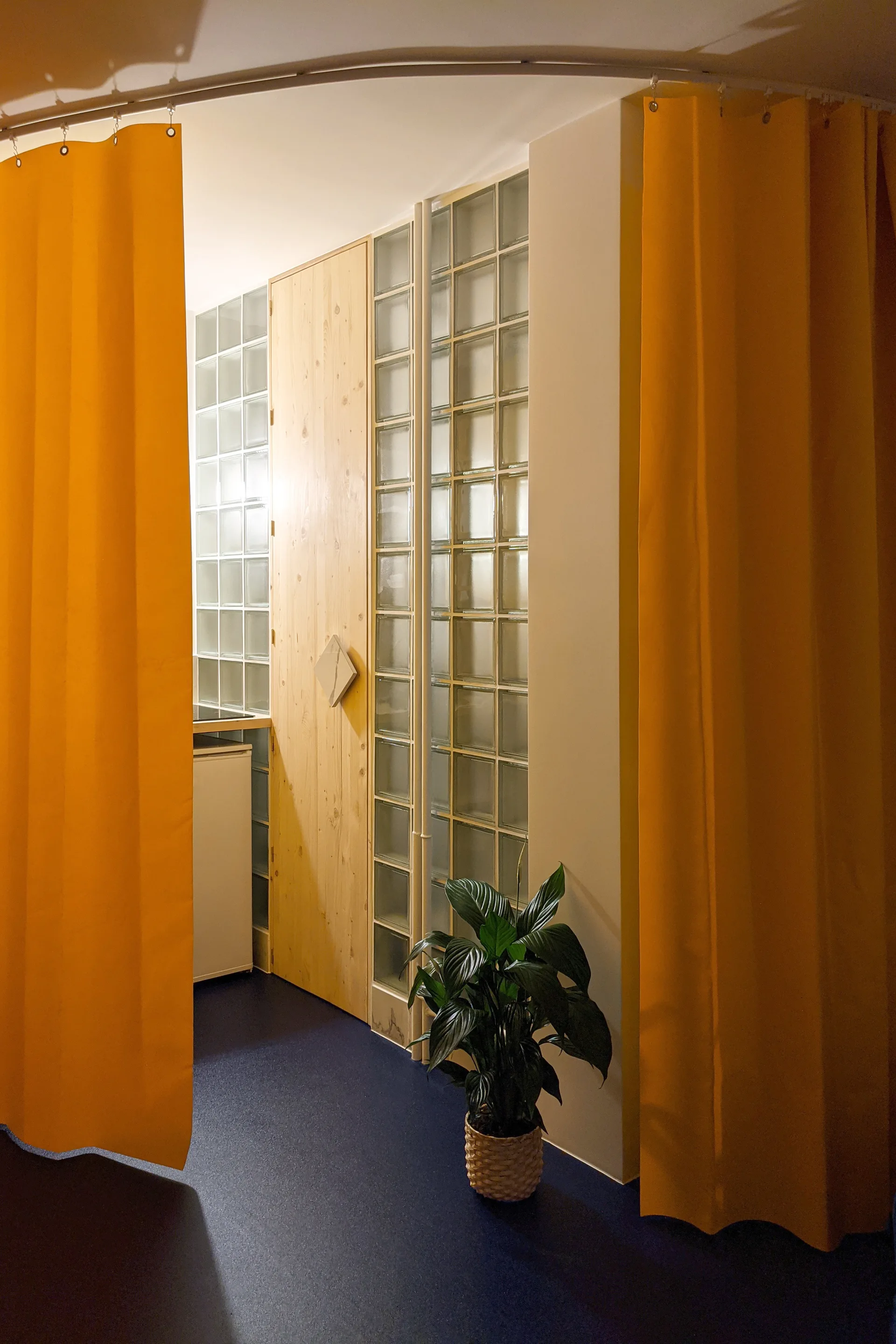 A modern interior space featuring a translucent glass block wall, a natural wood door, and vibrant yellow curtains on a curved ceiling track.