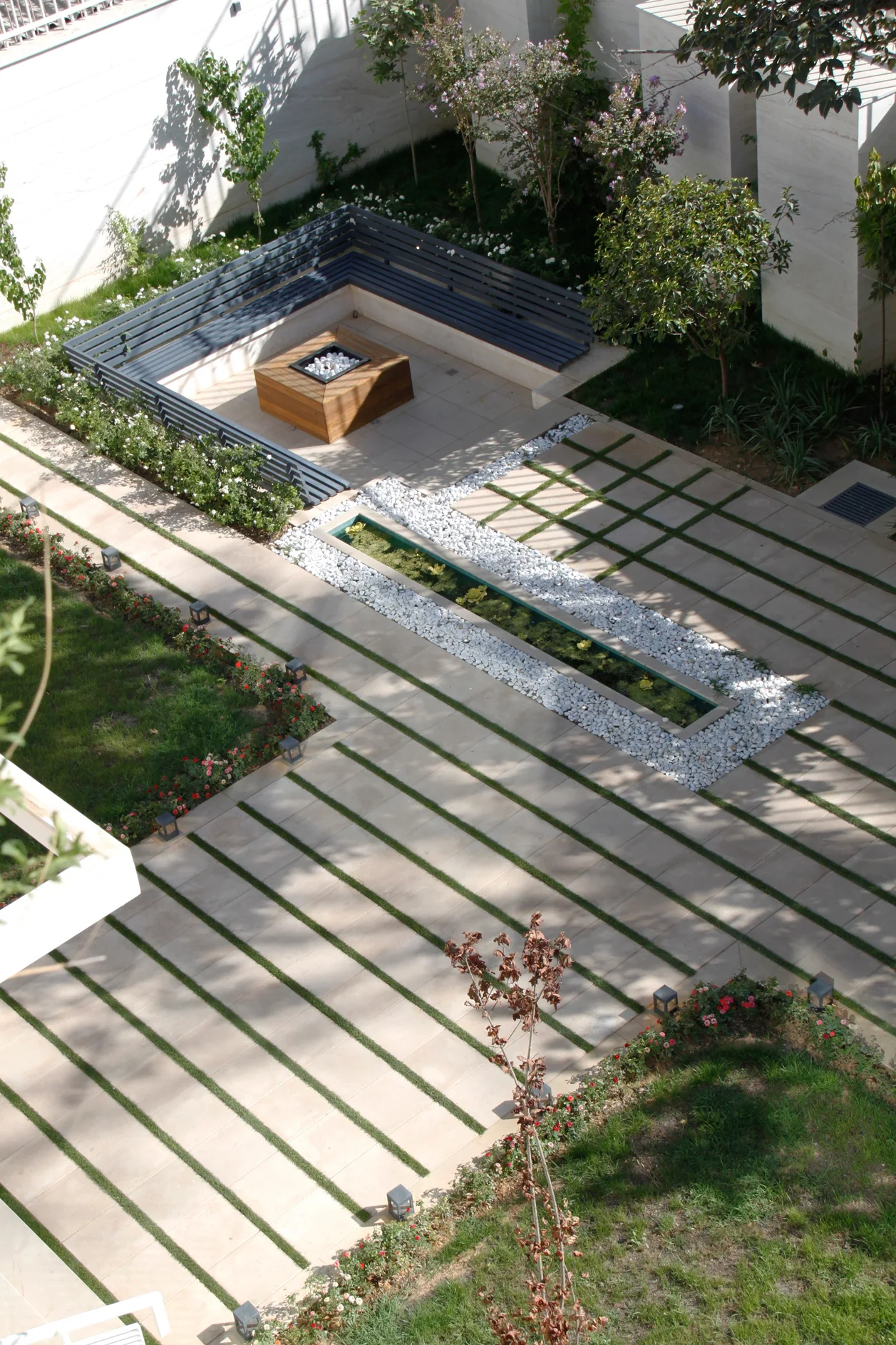 An overhead view of a modern landscaped garden with geometric stone pavers, a wooden fire pit surrounded by a dark bench, and a water feature.