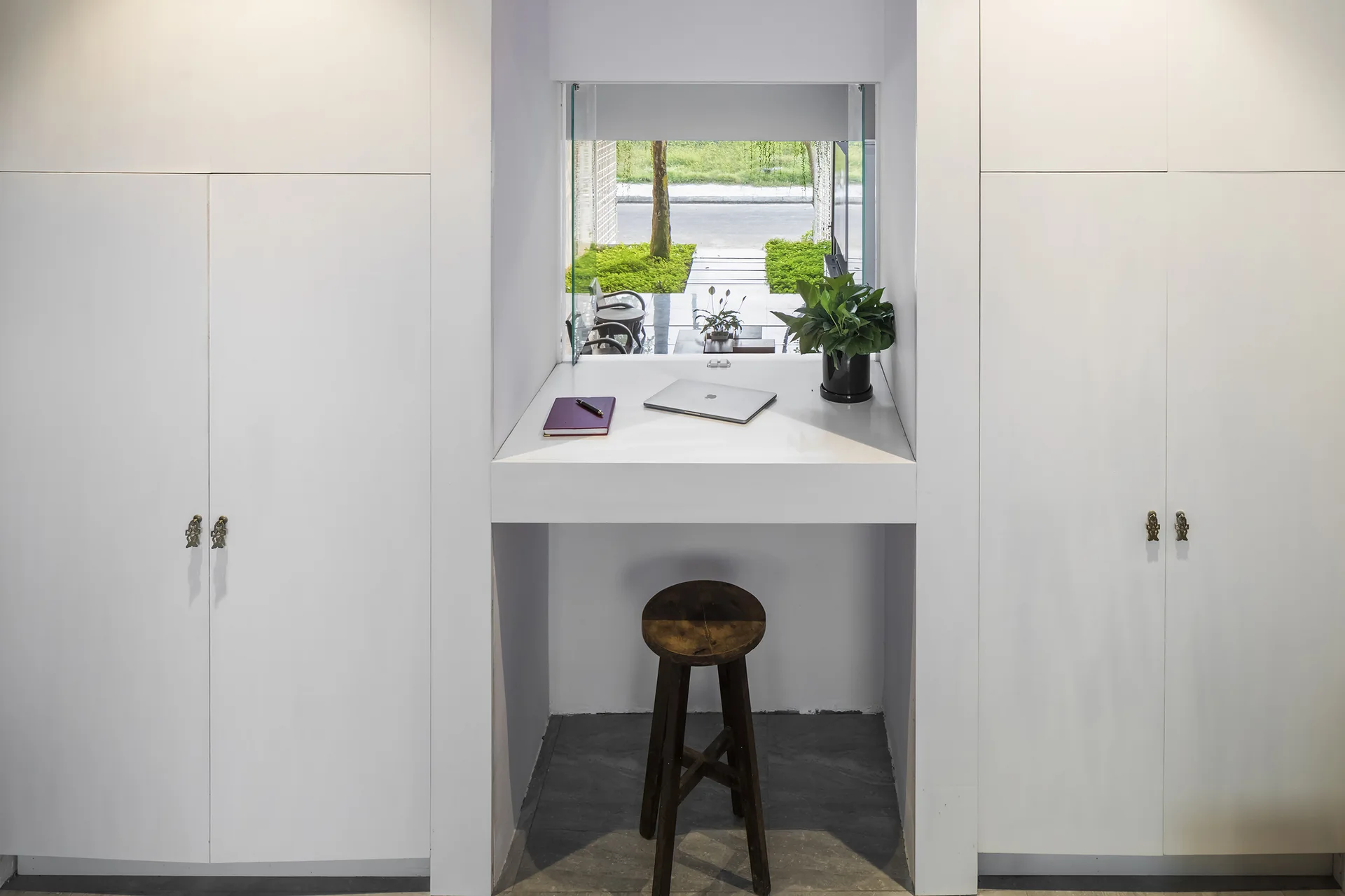 A minimalist white built-in desk with a rustic wooden stool sits beneath a low window overlooking a garden path.