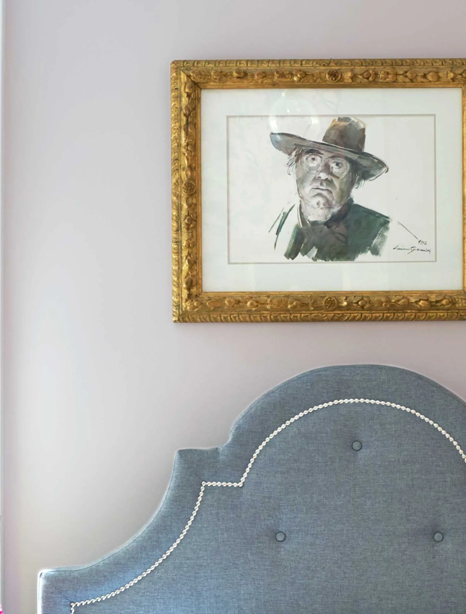 A close-up of a blue scalloped headboard with nailhead trim underneath an ornate gold-framed portrait on a lavender wall.