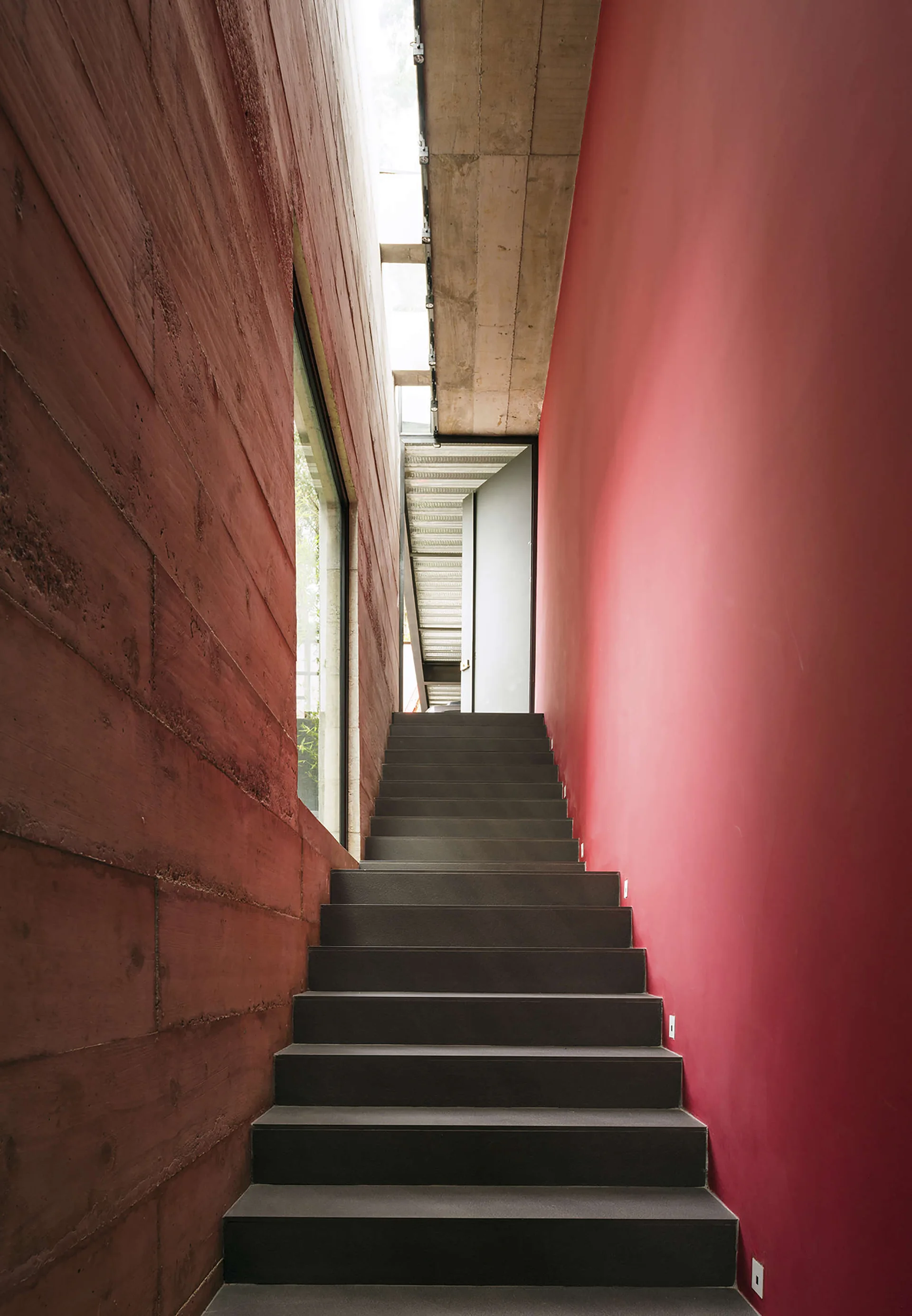 A dark grey concrete staircase rises between a textured reddish-brown concrete wall and a smooth painted red wall beneath a narrow skylight.