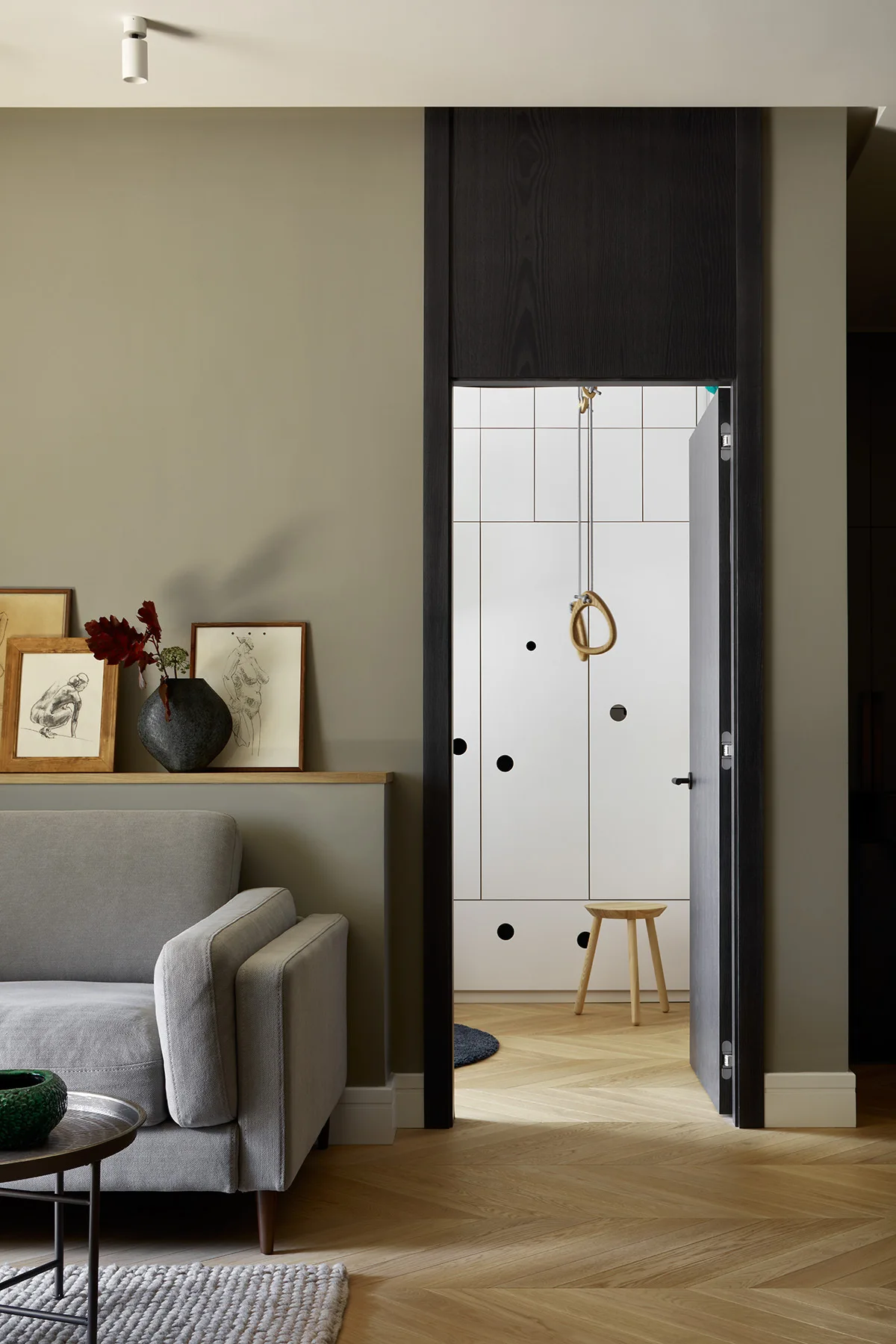 A grey sofa sits against a sage green wall with a shallow display ledge next to a black-framed doorway showing an adjoining room with gymnastics rings.