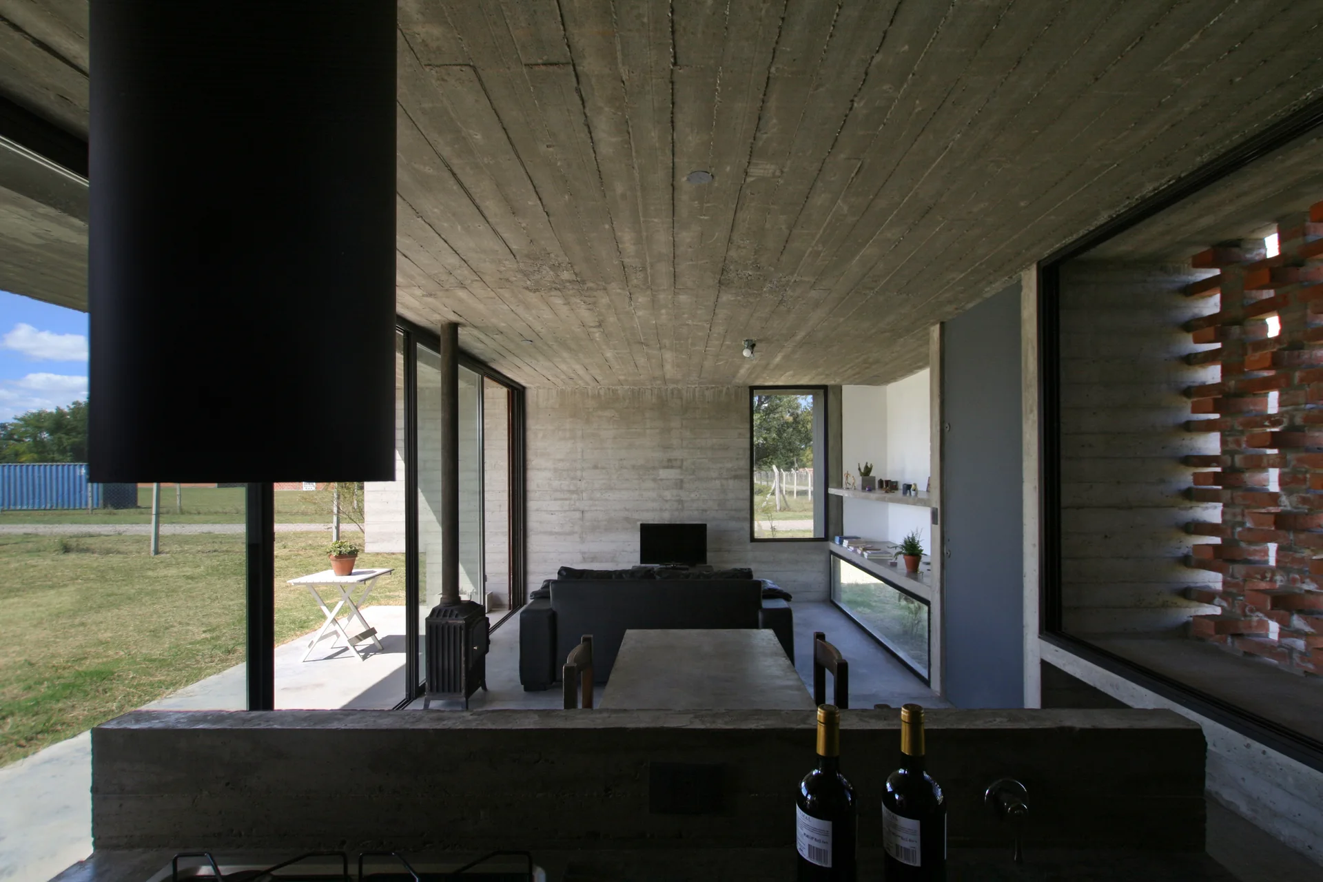 A modern living and dining room featuring raw concrete walls and ceiling, a dark sofa, a freestanding metal fireplace, and a decorative brick screen wall.