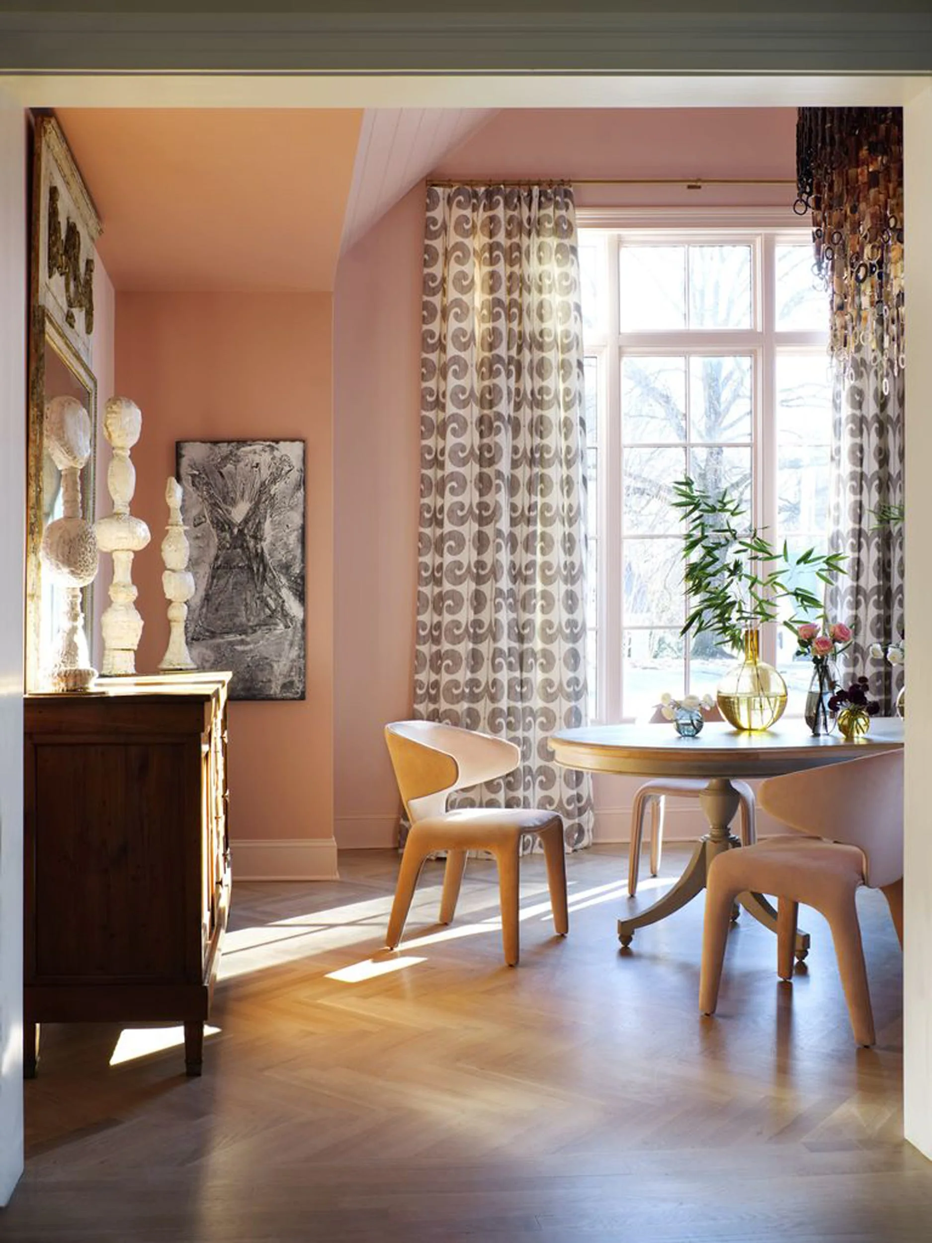 A bright dining room features blush pink walls, sculptural wooden chairs around a round table, and patterned floor-length curtains by a large window.