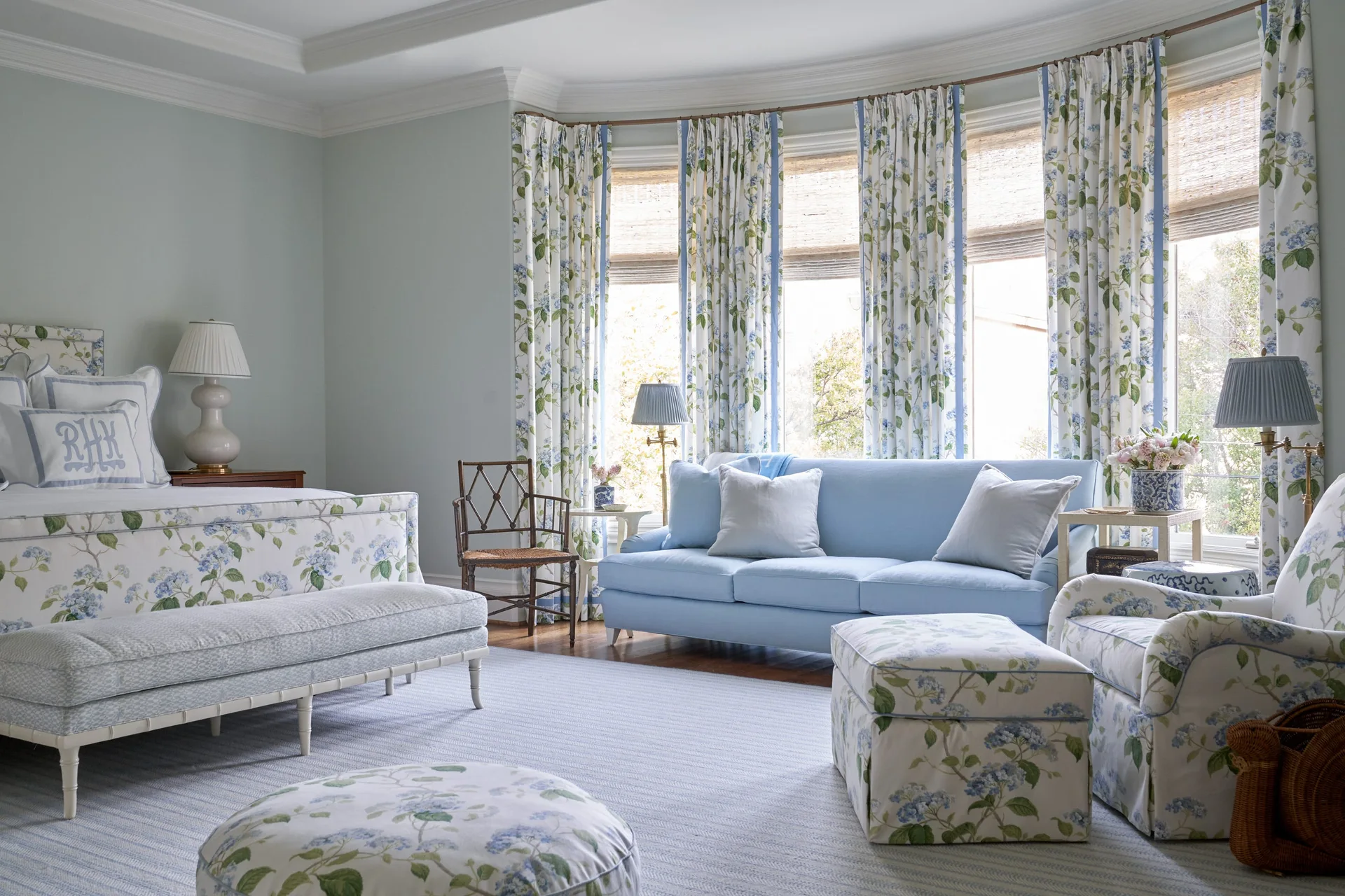 A traditional light blue bedroom features a bed with floral bedding, matching curtains, an armchair, and a solid blue sofa in a bay window.