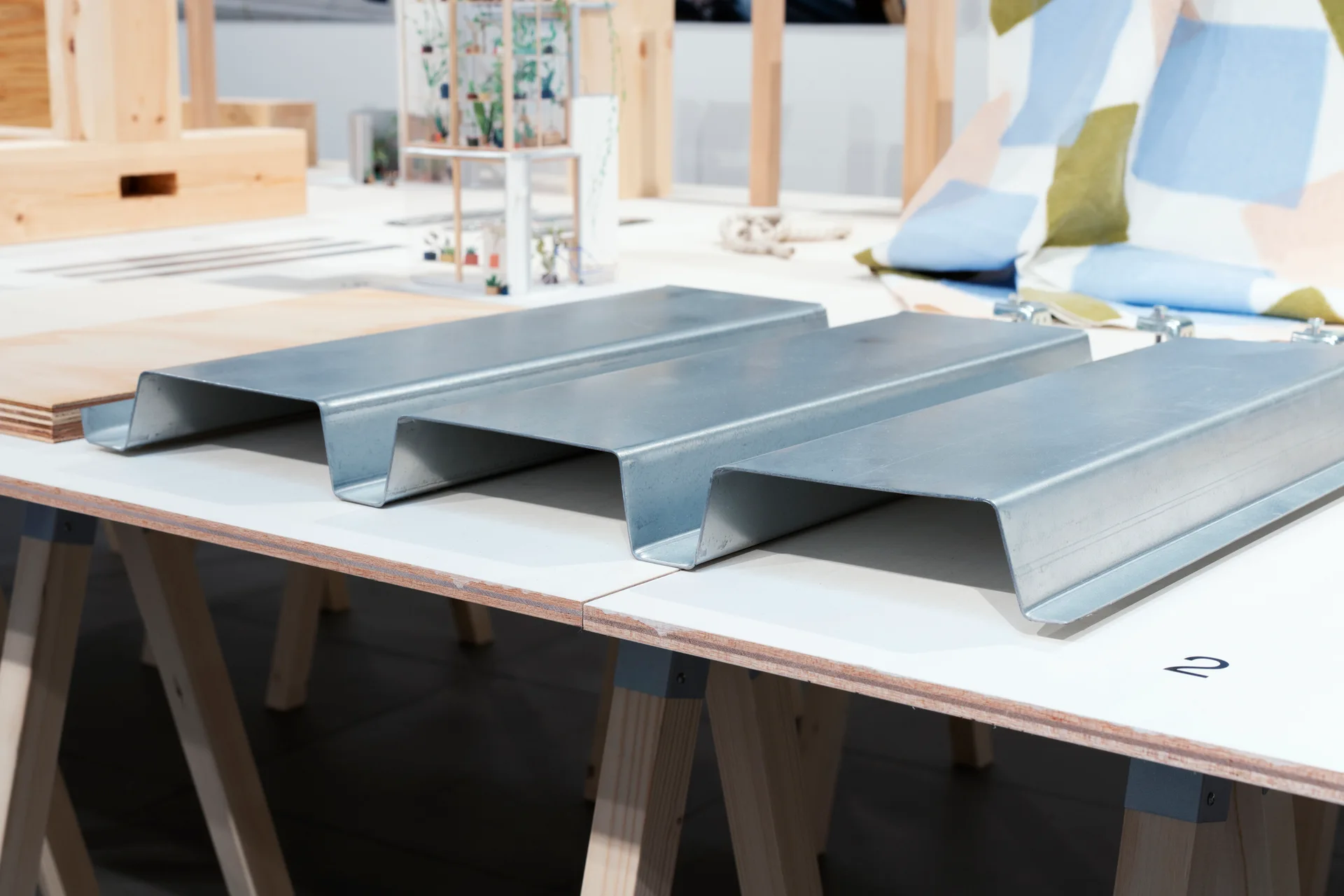 A corrugated galvanized metal profile sample lies on a white wooden workbench with sawhorses in an architectural design studio.