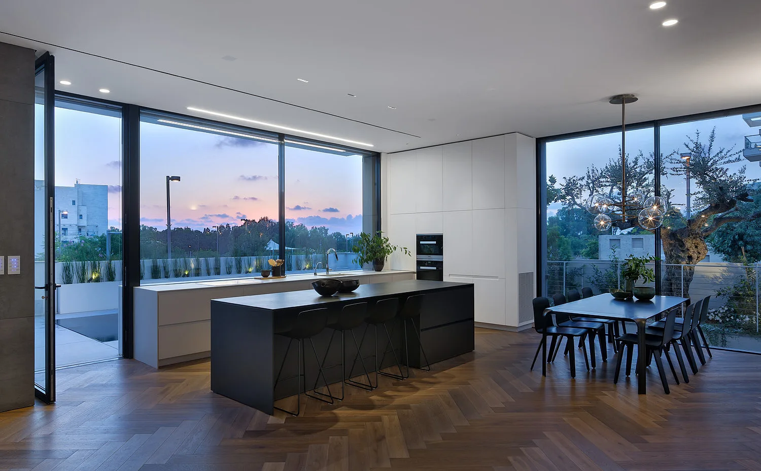 A modern open-plan kitchen featuring a large black island, white built-in cabinetry, and herringbone wood flooring next to large glass windows.