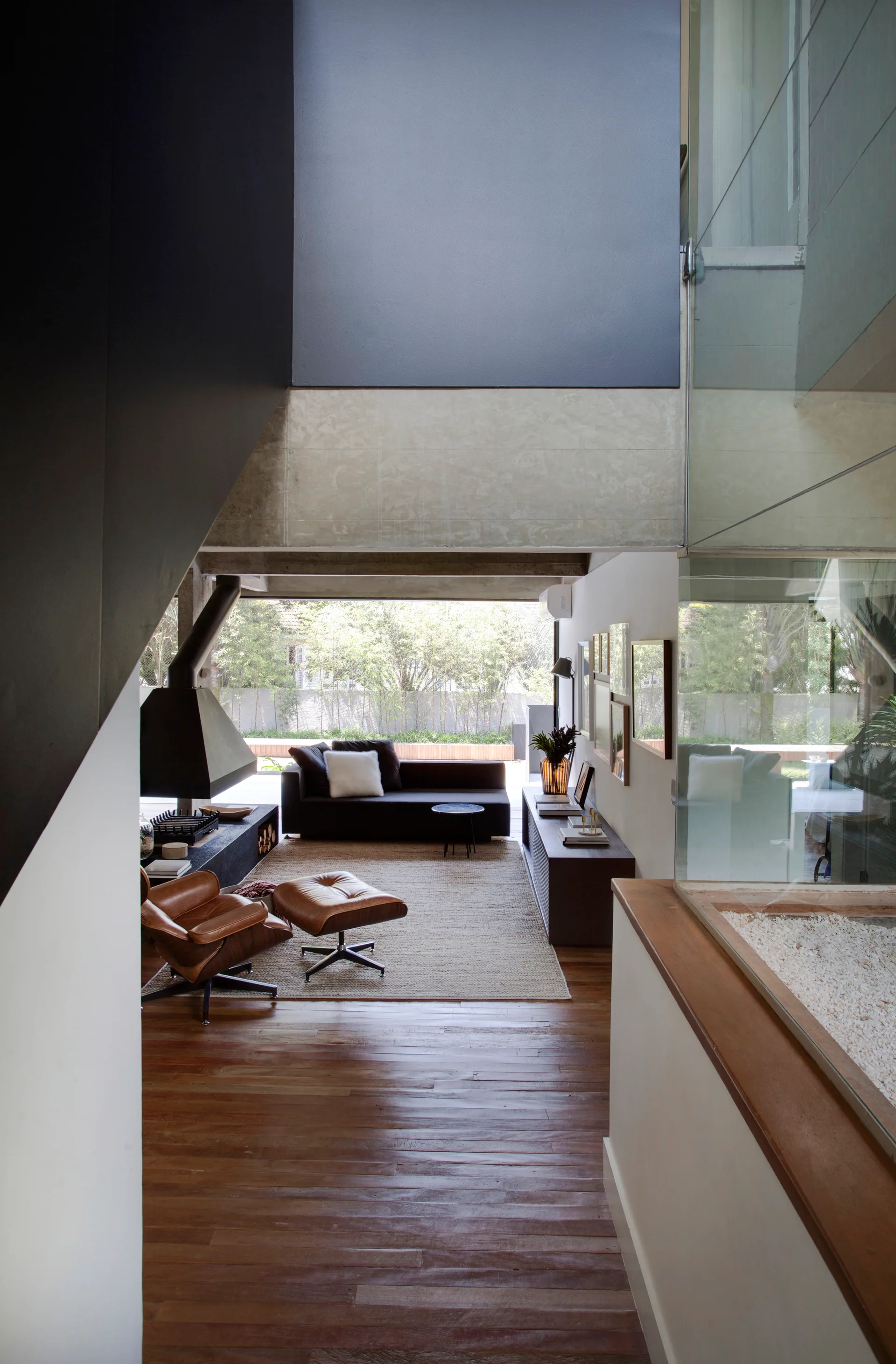 A split-level view of a modern living room featuring a suspended black fireplace, a leather lounge chair, and a large window looking onto a garden.