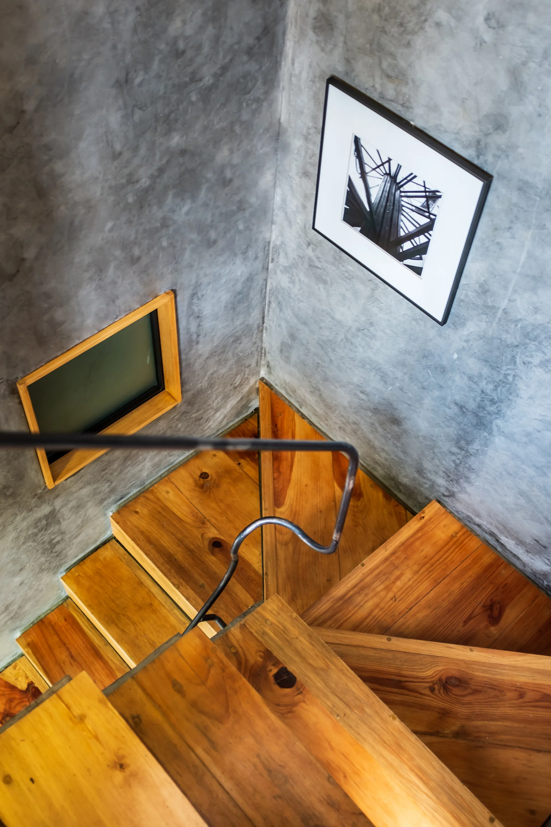Looking down a winding wood staircase surrounded by textured grey concrete walls, featuring a thin black metal railing and framed art.