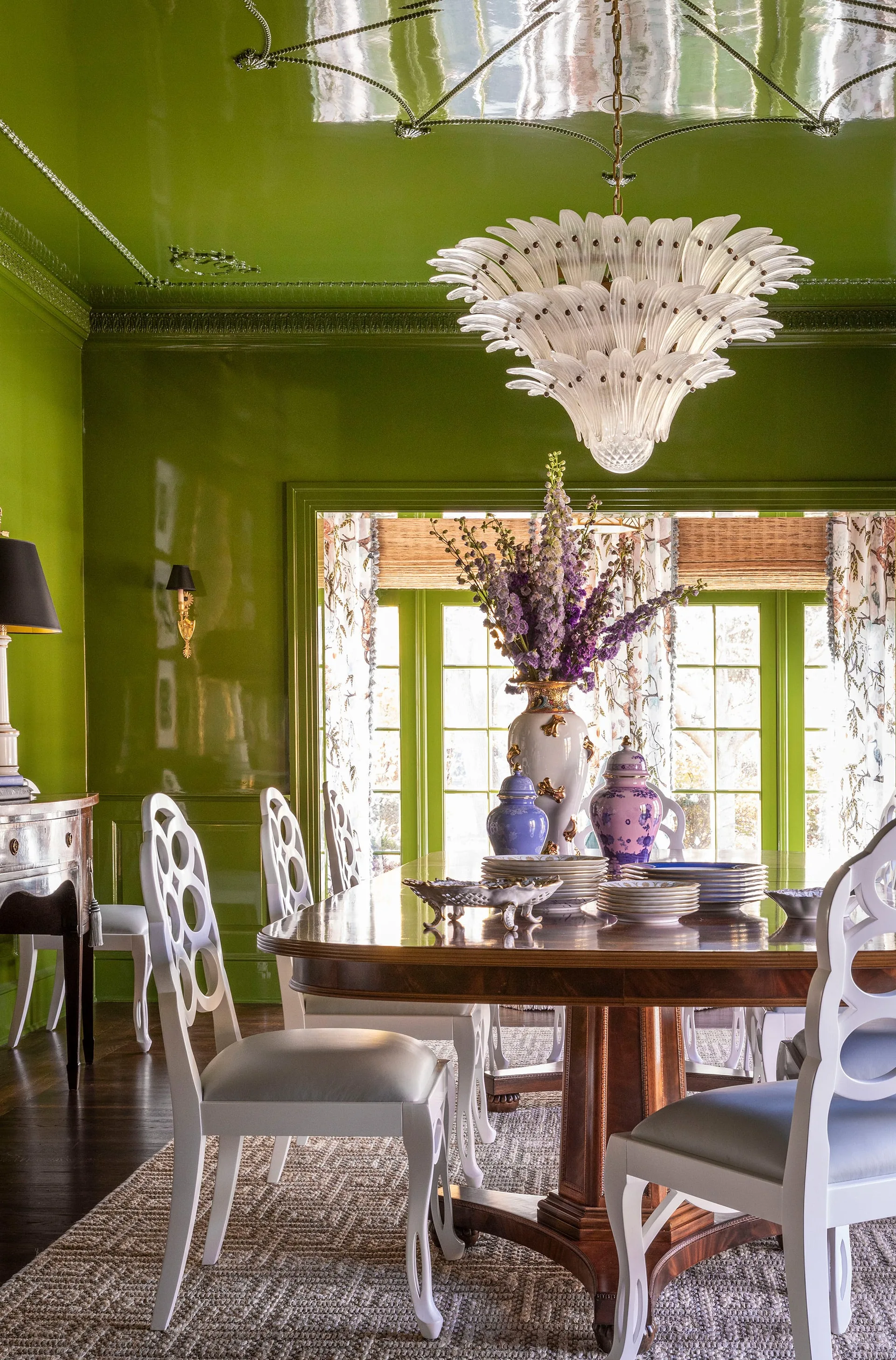 A traditional dining room features reflective green lacquered walls, a dark wood table with white cutout-back chairs, and a glass floral chandelier.
