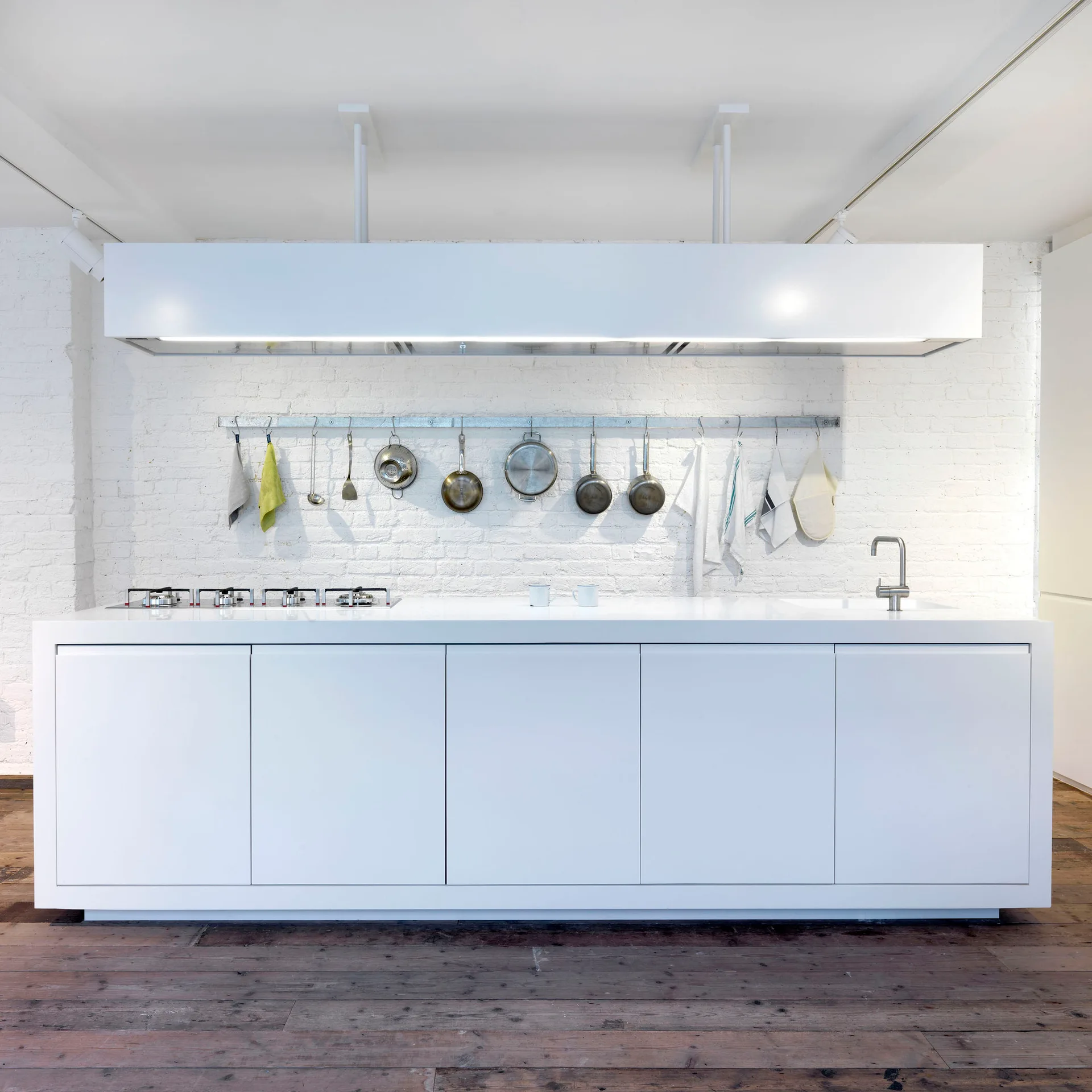 A minimalist white kitchen island with a sink and stovetop sits in front of a white painted brick wall with hanging metal cookware.
