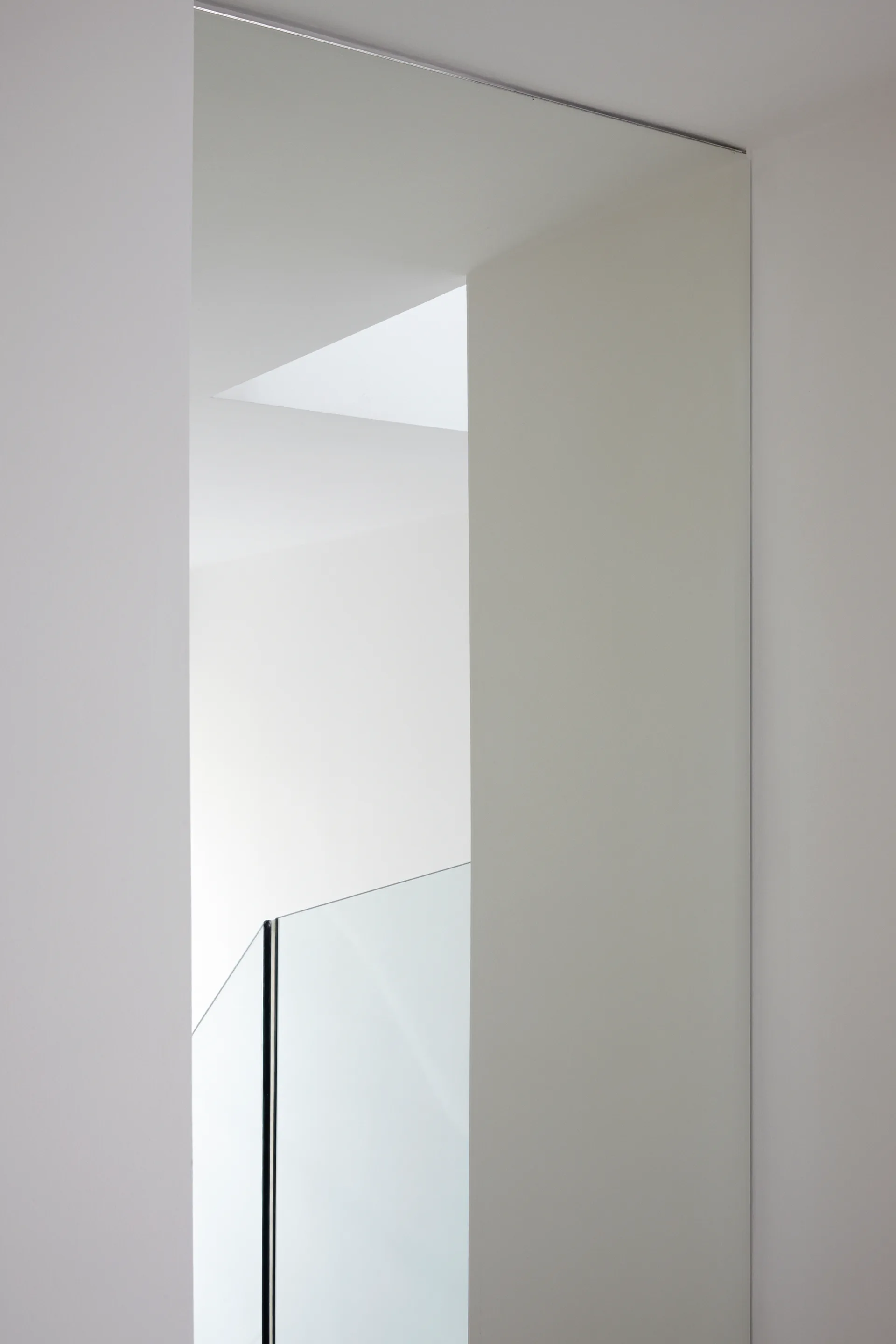 A minimalist white stairwell features intersecting wall planes, a transparent glass balustrade, and natural light from a square skylight.