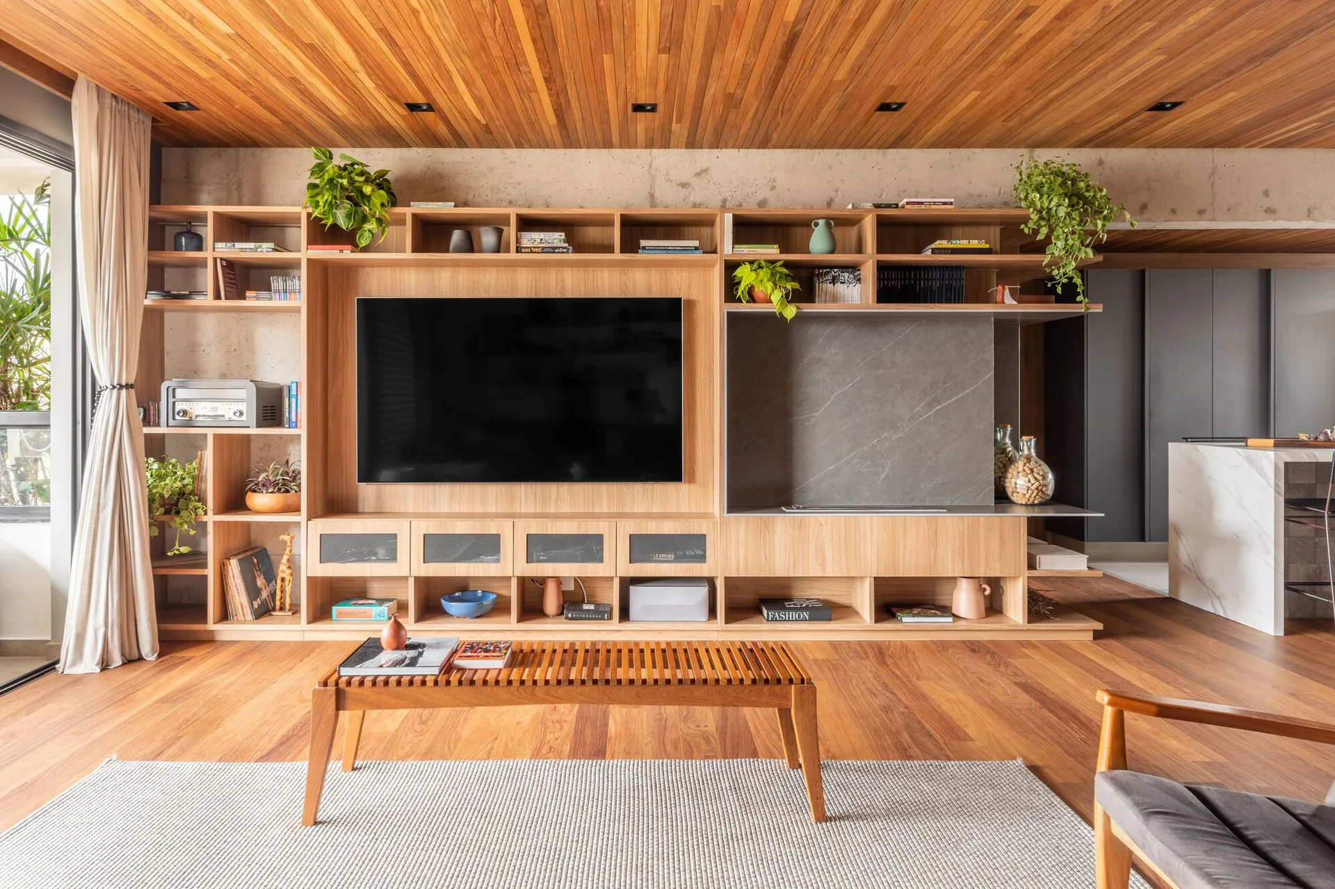 A modern living room features a custom wood media wall built-in below a slatted timber ceiling and a raw concrete accent band.