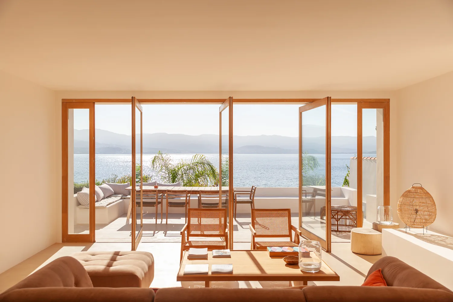 A sunlit living room with wood-framed folding doors open to a patio with a dining table and built-in bench overlooking the ocean.