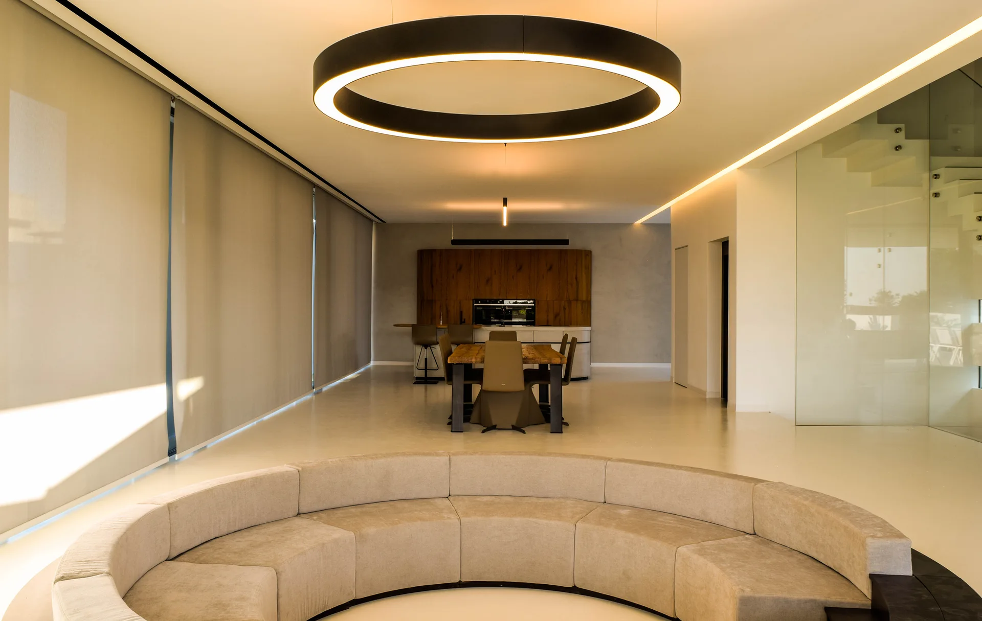 A large curved beige sectional sits in a spacious open-concept living area beneath a massive circular ring pendant light.