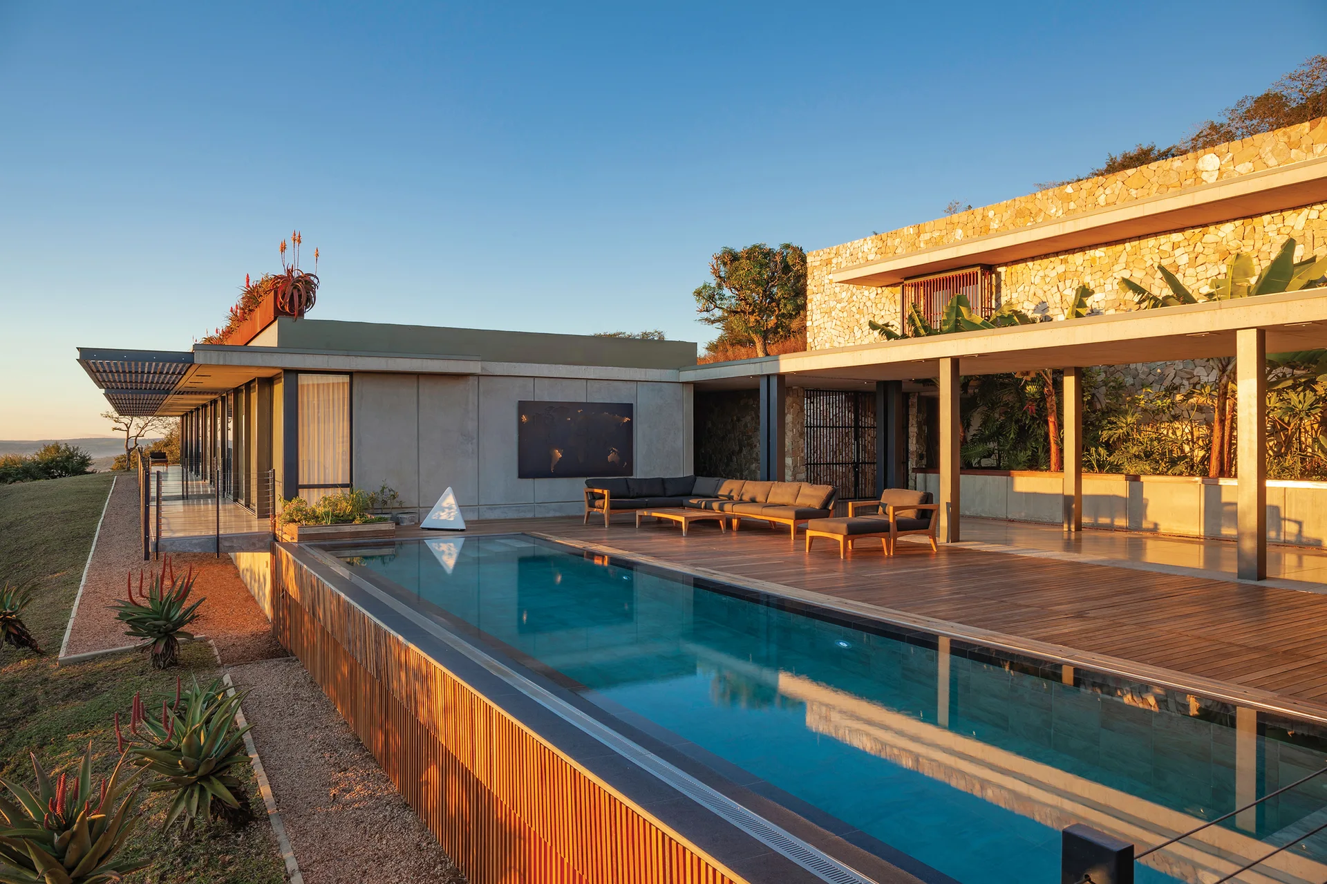 A rectangular infinity pool borders a spacious wooden deck with a modular grey sofa set against a natural stone wall.