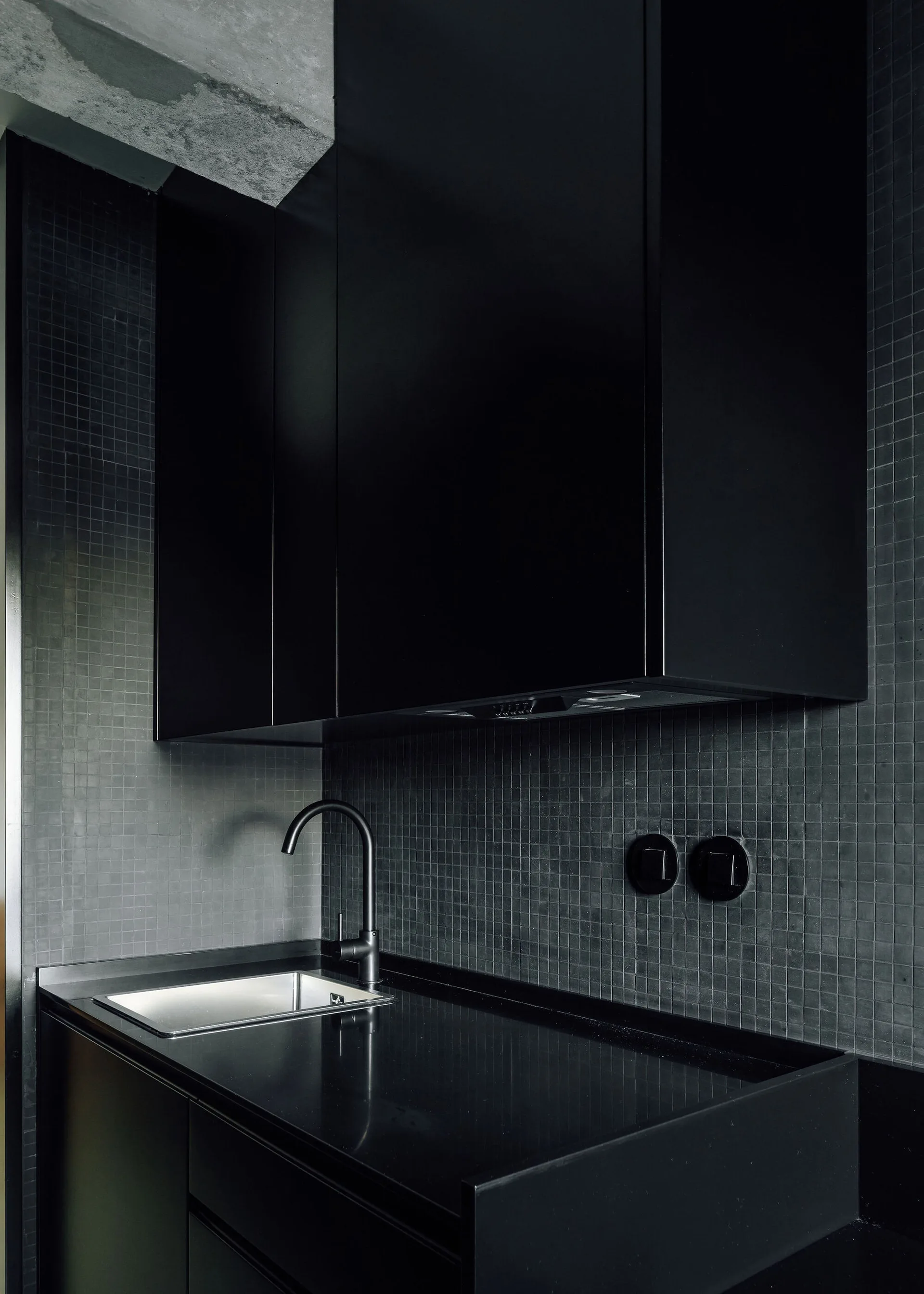 A modern dark kitchen features matte black handleless cabinets, a matching countertop, and a dark grey tiled backsplash in a small grid pattern.