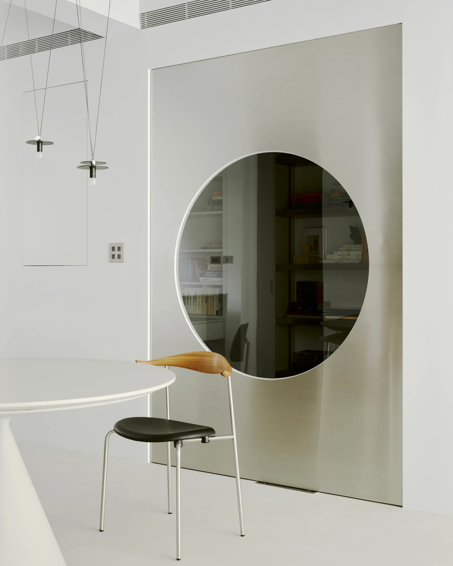 A minimalist dining space features a white round table, a modern chair, and a brushed metal sliding door with a large circular window opening.