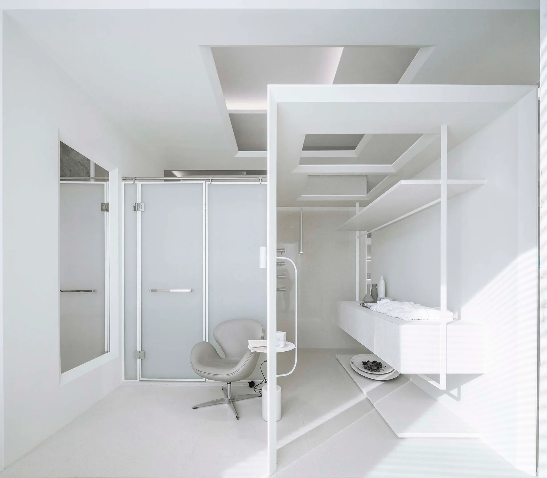A monochromatic white interior featuring a gray swivel armchair, a frosted glass sliding partition, and custom geometric architectural shelving.