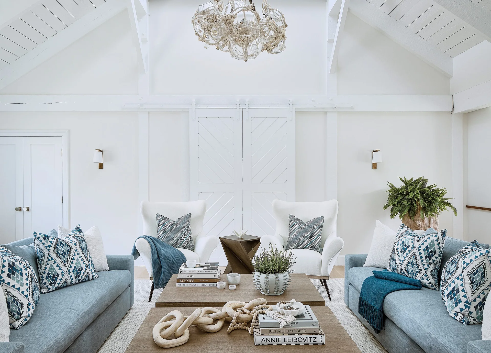 A symmetrical living room featuring two blue sofas and two white armchairs arranged around a wooden coffee table in front of white barn doors.