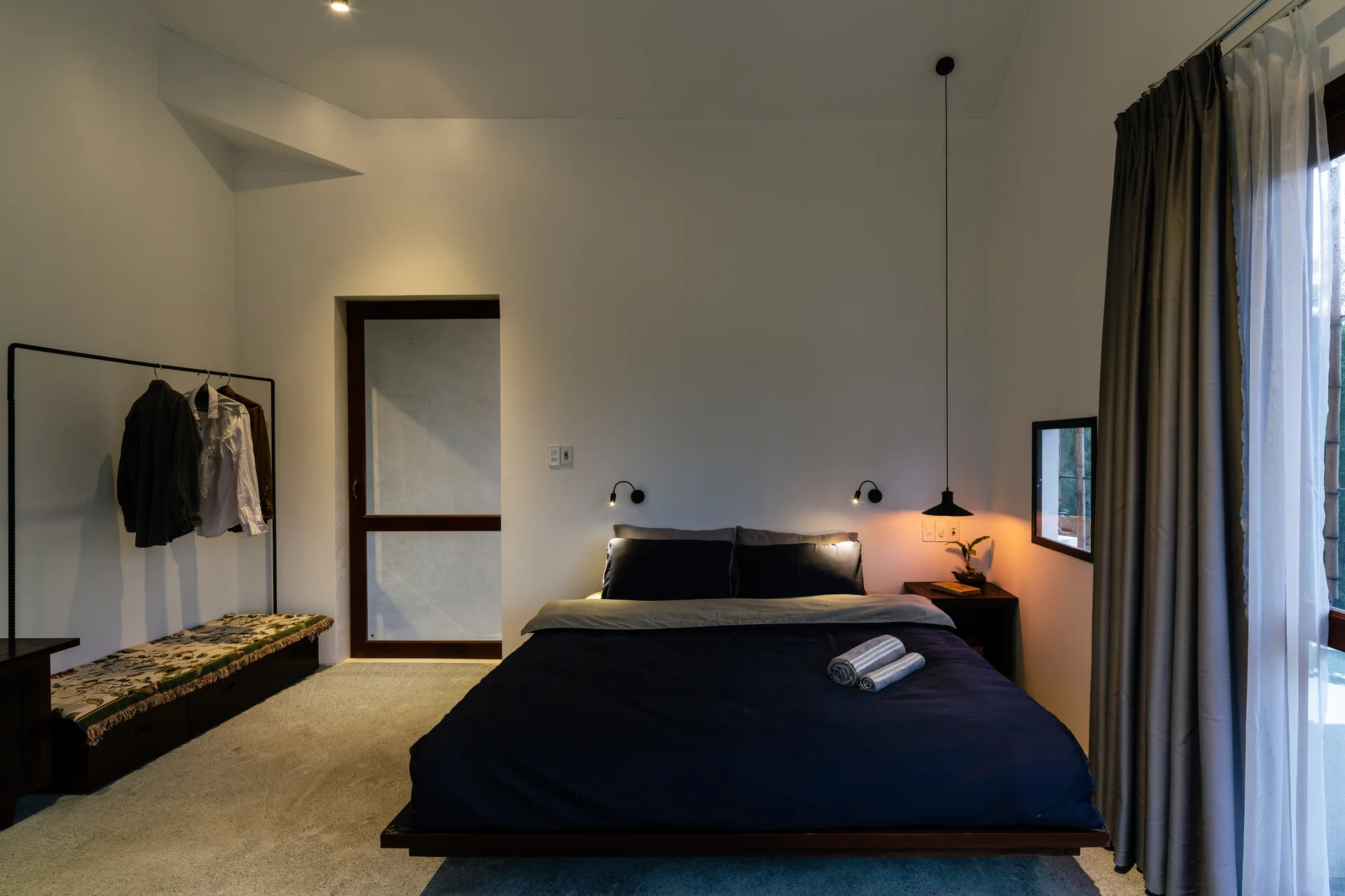 A minimalist bedroom features a navy platform bed, a black metal garment rack, and warm hanging pendant lighting against white walls.