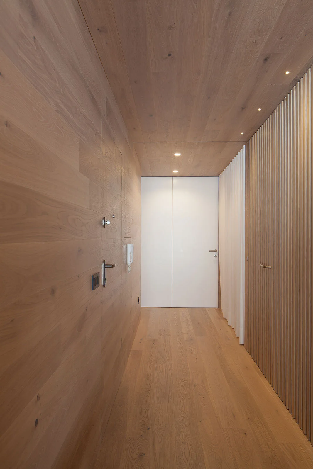 A narrow hallway features warm wood paneling on the floor, walls, and ceiling, ending at a minimalist white door.