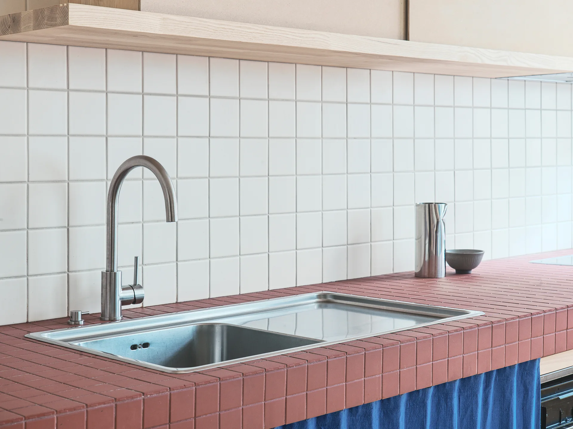 A stainless steel sink is set into a red tiled countertop with a white square tiled backsplash, a light wood shelf, and a blue under-sink curtain.