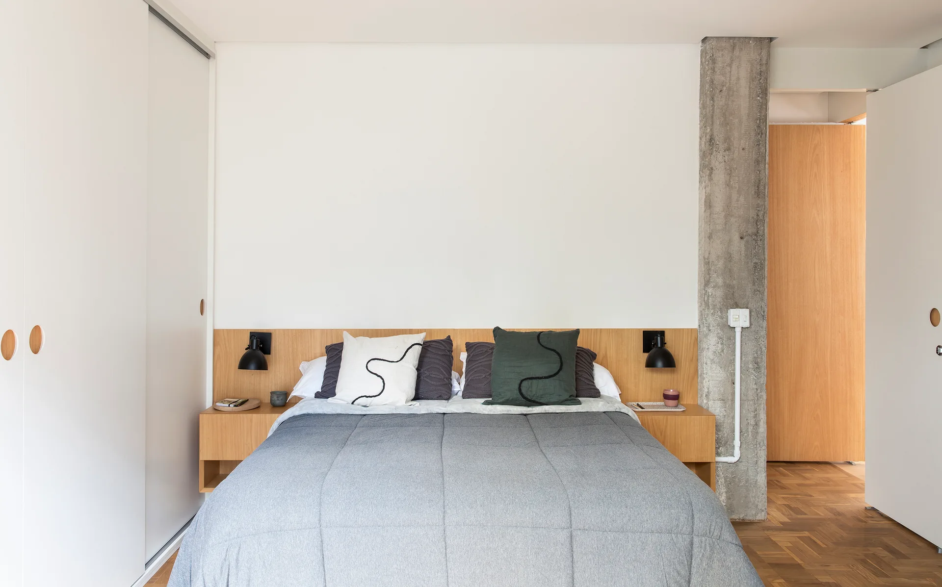 A minimalist bedroom featuring a bed with an integrated wood headboard and nightstands, next to an exposed concrete column and parquet floor.