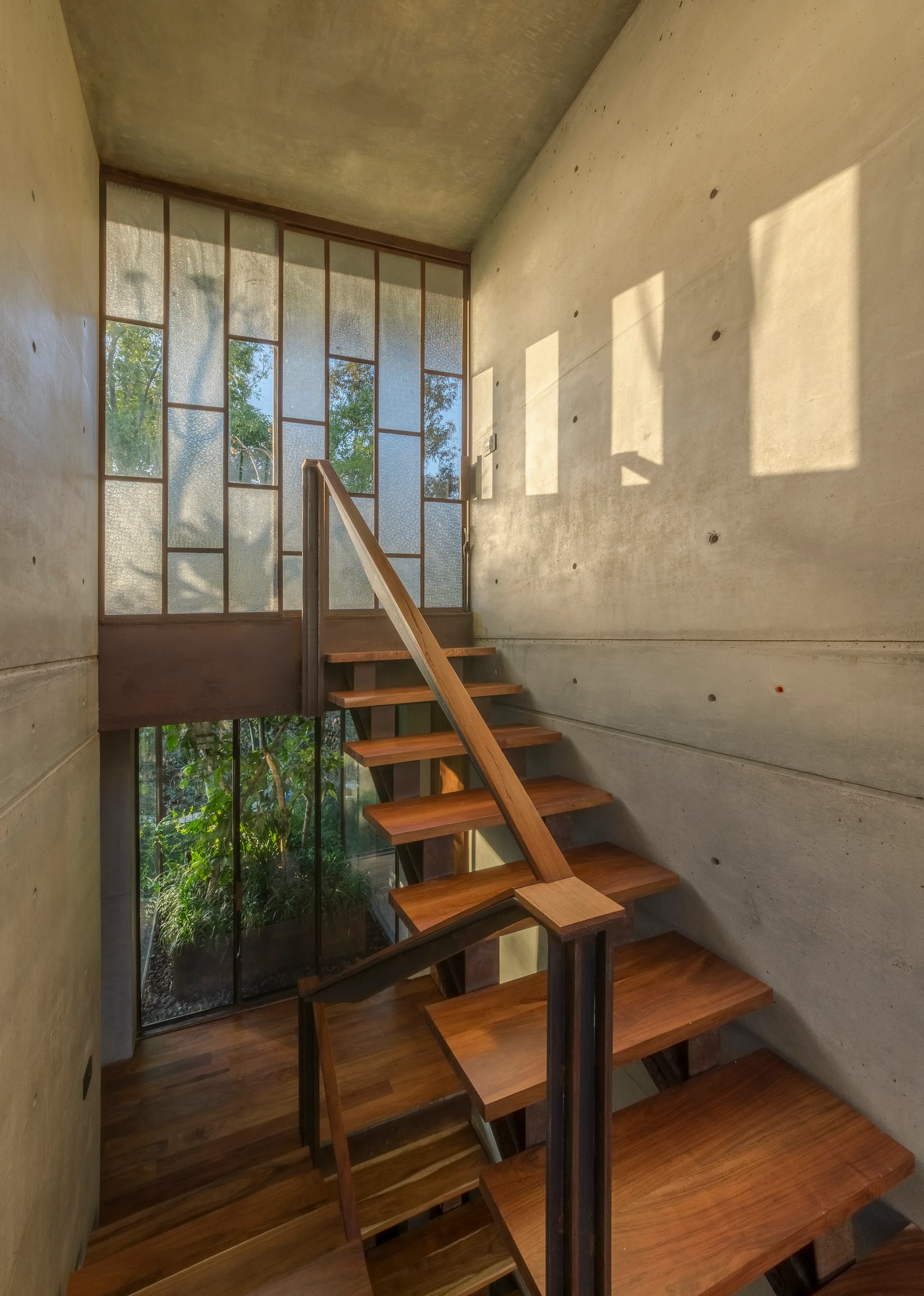 A wooden staircase with floating treads climbs between raw concrete walls next to a geometric textured glass screen in natural sunlight.