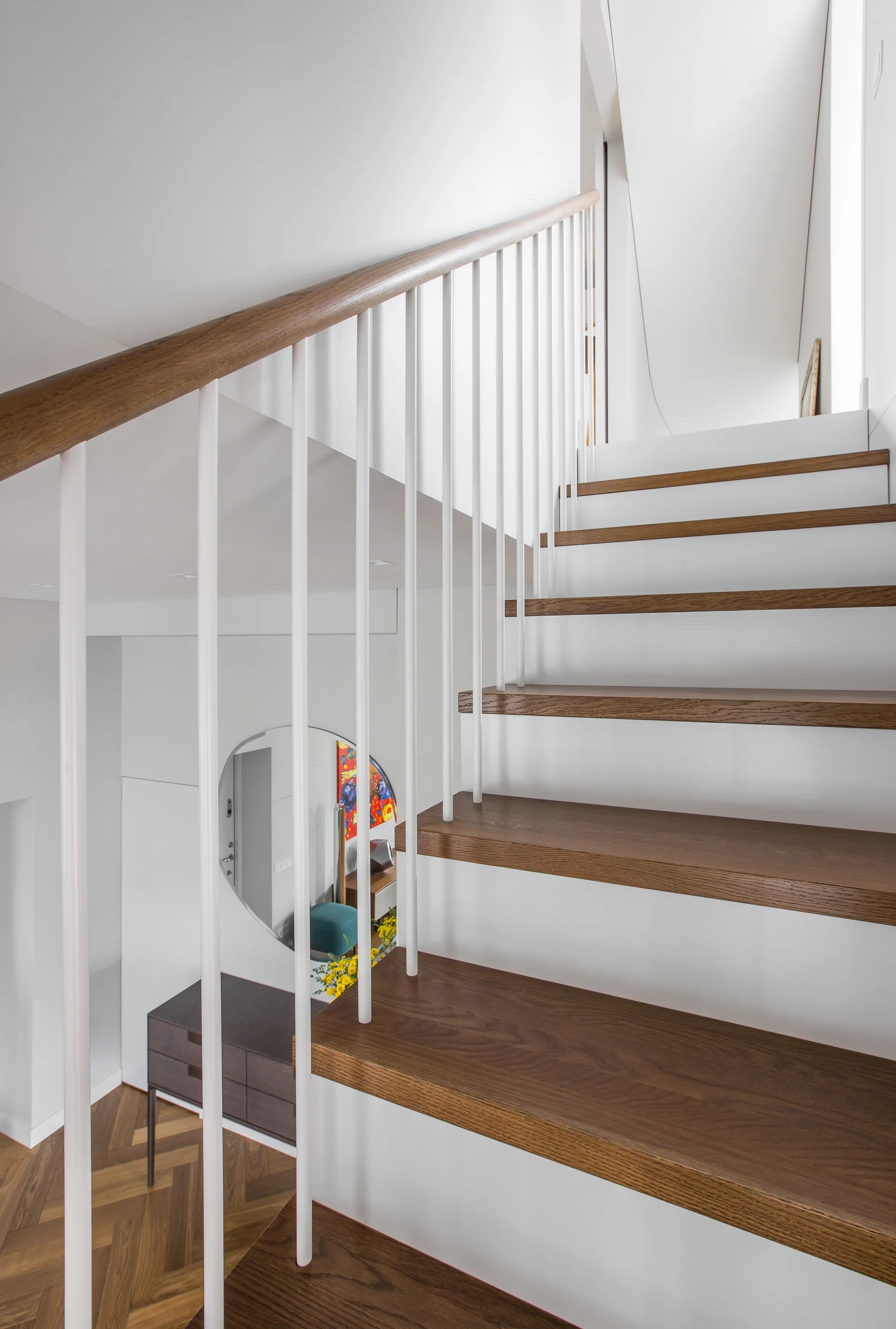 A modern staircase features dark wood treads, white risers, and thin white spindles, looking down toward a herringbone wood floor and round entryway mirror.