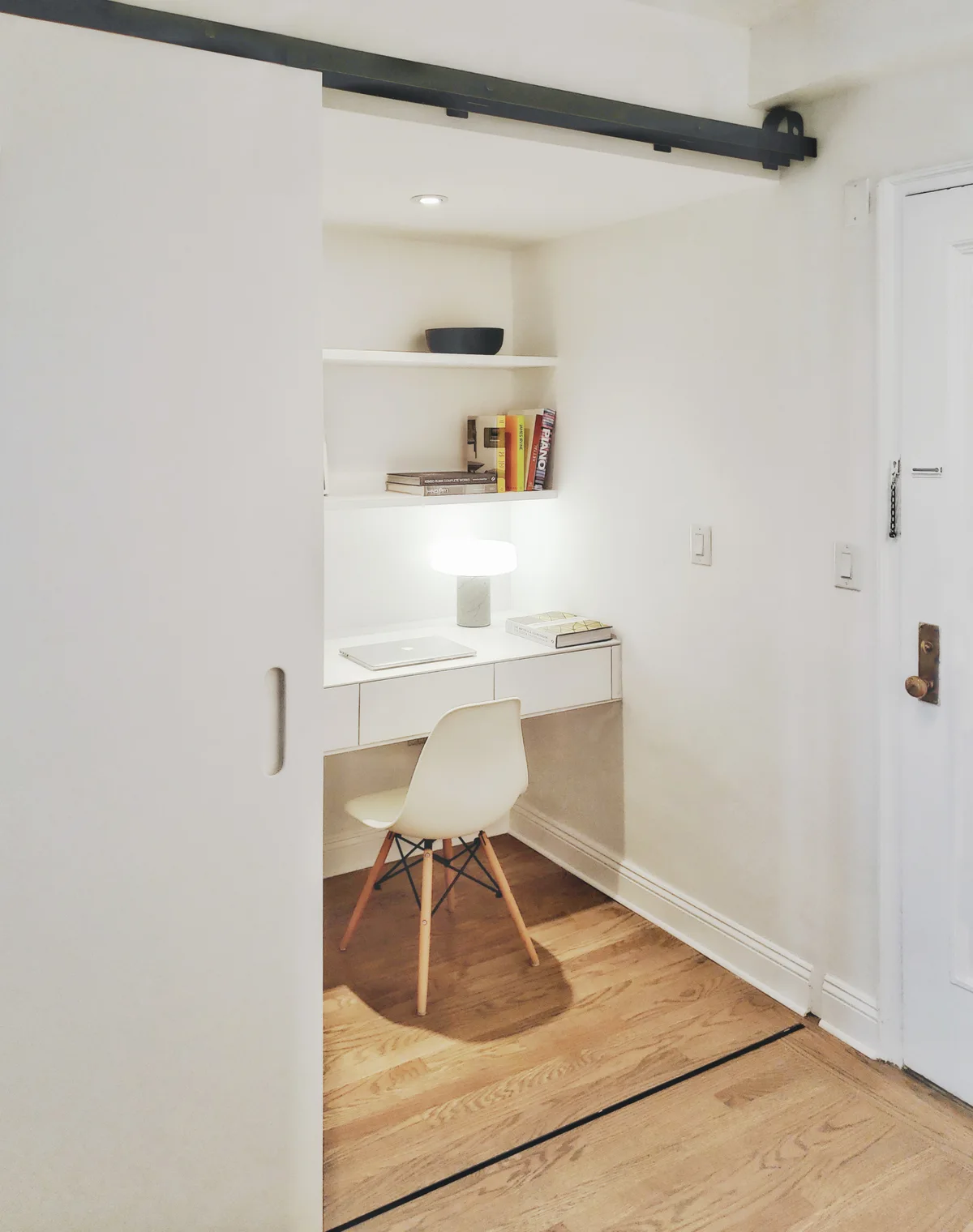 A compact home office nook with a floating white desk and chair set inside an alcove behind a white sliding door.