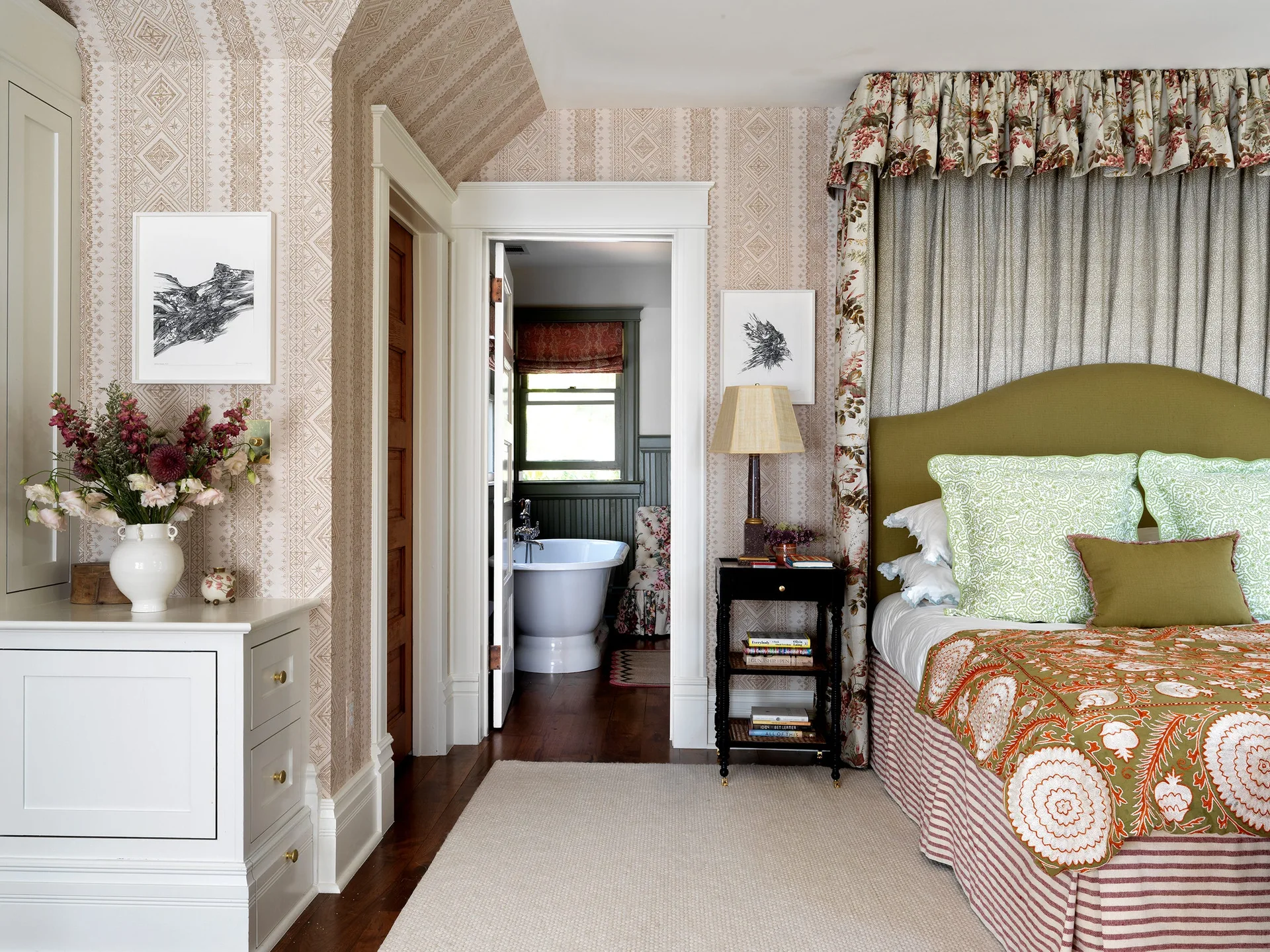 A bedroom with patterned wallpaper features an olive green headboard and floral canopy next to an open doorway showing a white bathtub.