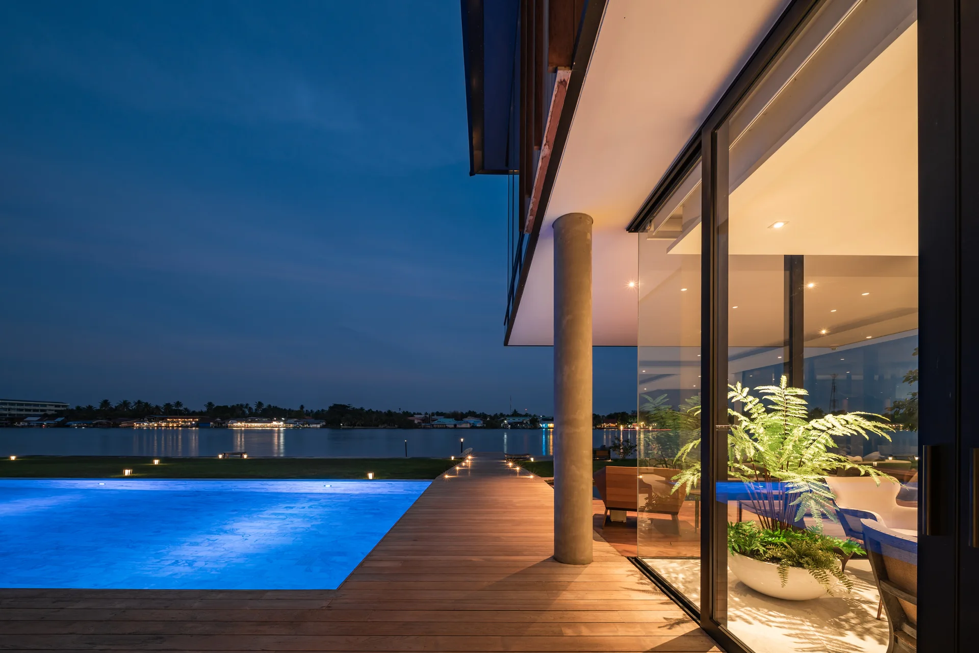 An illuminated swimming pool glows blue next to a wooden deck and a modern home with glass walls at dusk, overlooking a river.