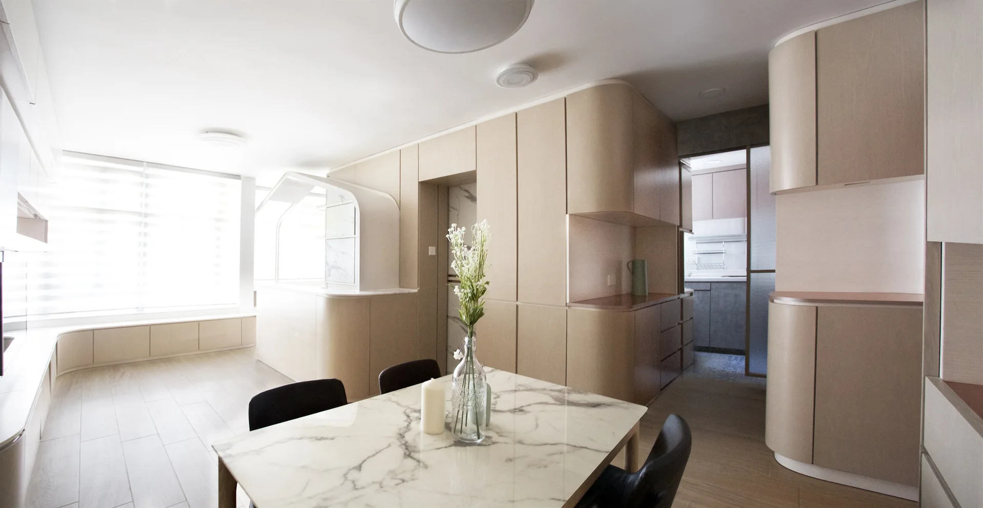 A minimalist dining space featuring a veined marble table, black chairs, and extensive curved light wood cabinetry under soft ceiling lights.