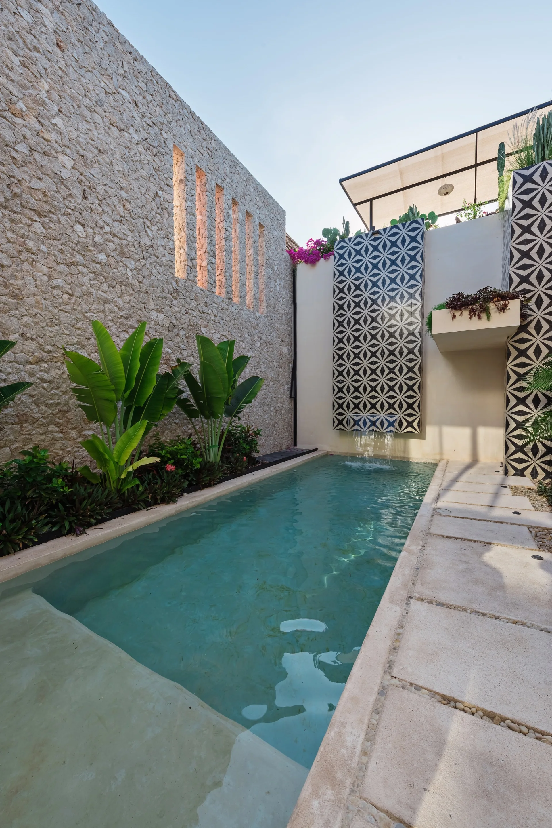 An outdoor rectangular swimming pool with a high-contrast geometric tiled waterfall and a textured stone wall with vertical slot cutouts.
