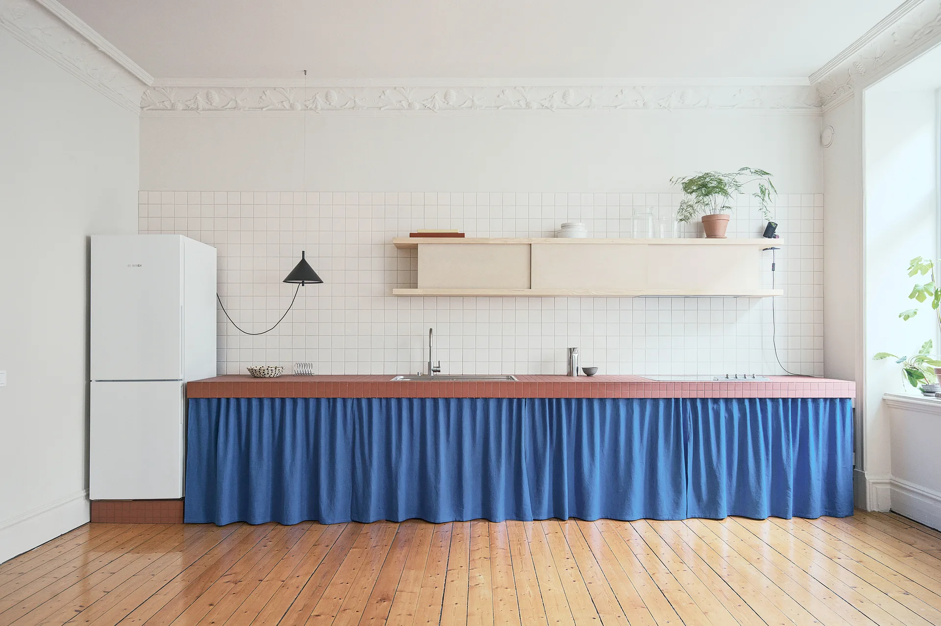 A minimalist kitchen features a long counter topped with terracotta tiles and covered by a gathered blue fabric skirt beneath a white tiled backsplash.