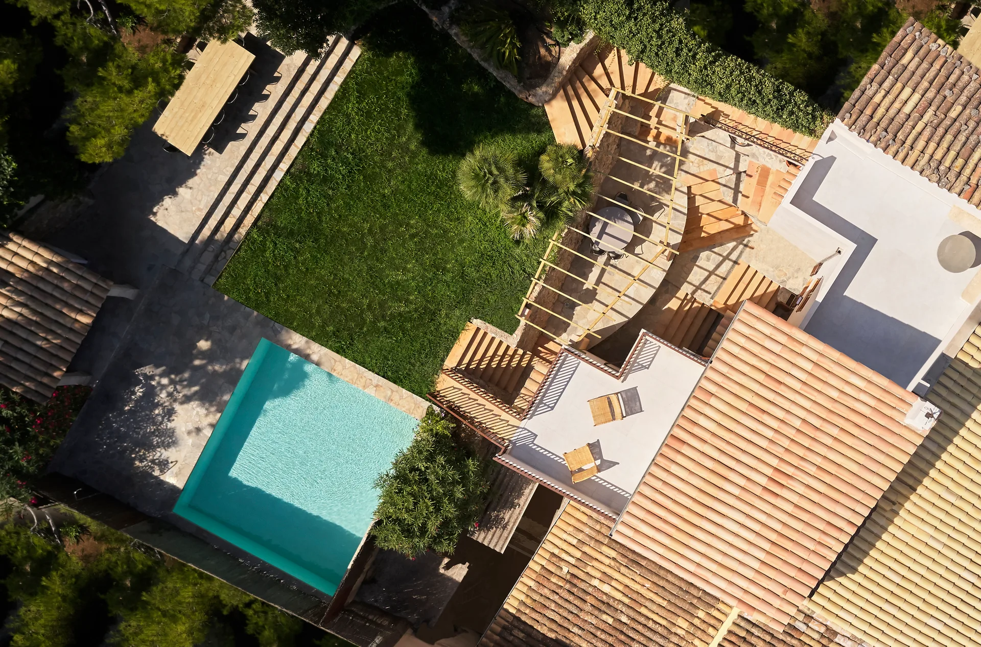 An aerial view of a multi-level Mediterranean backyard featuring a turquoise swimming pool, stone patios, a green lawn, and terracotta-tiled roofs.