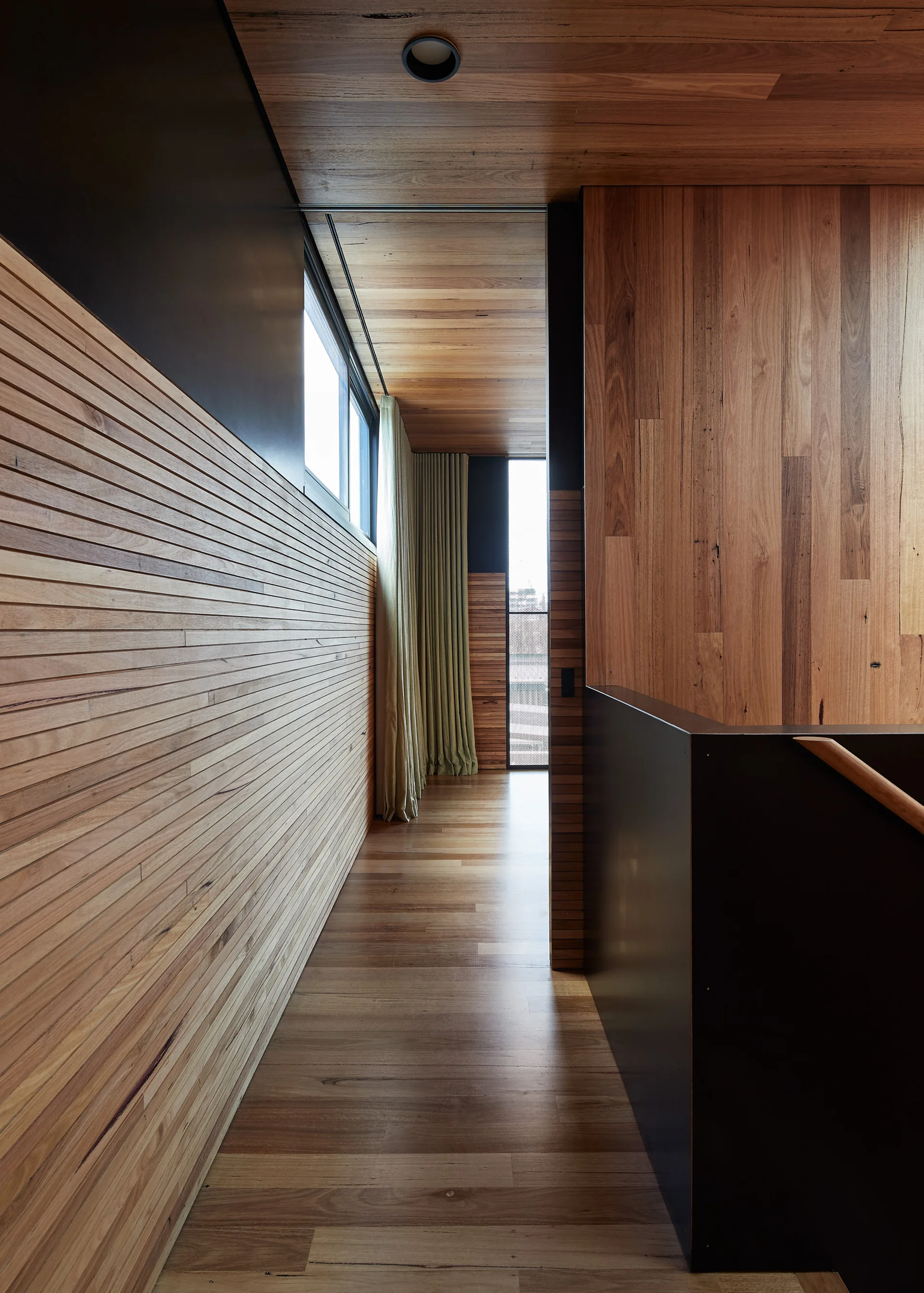 A modern hallway features horizontal slatted wood wall paneling, matching timber floors and ceiling, a dark partition, and a long window.