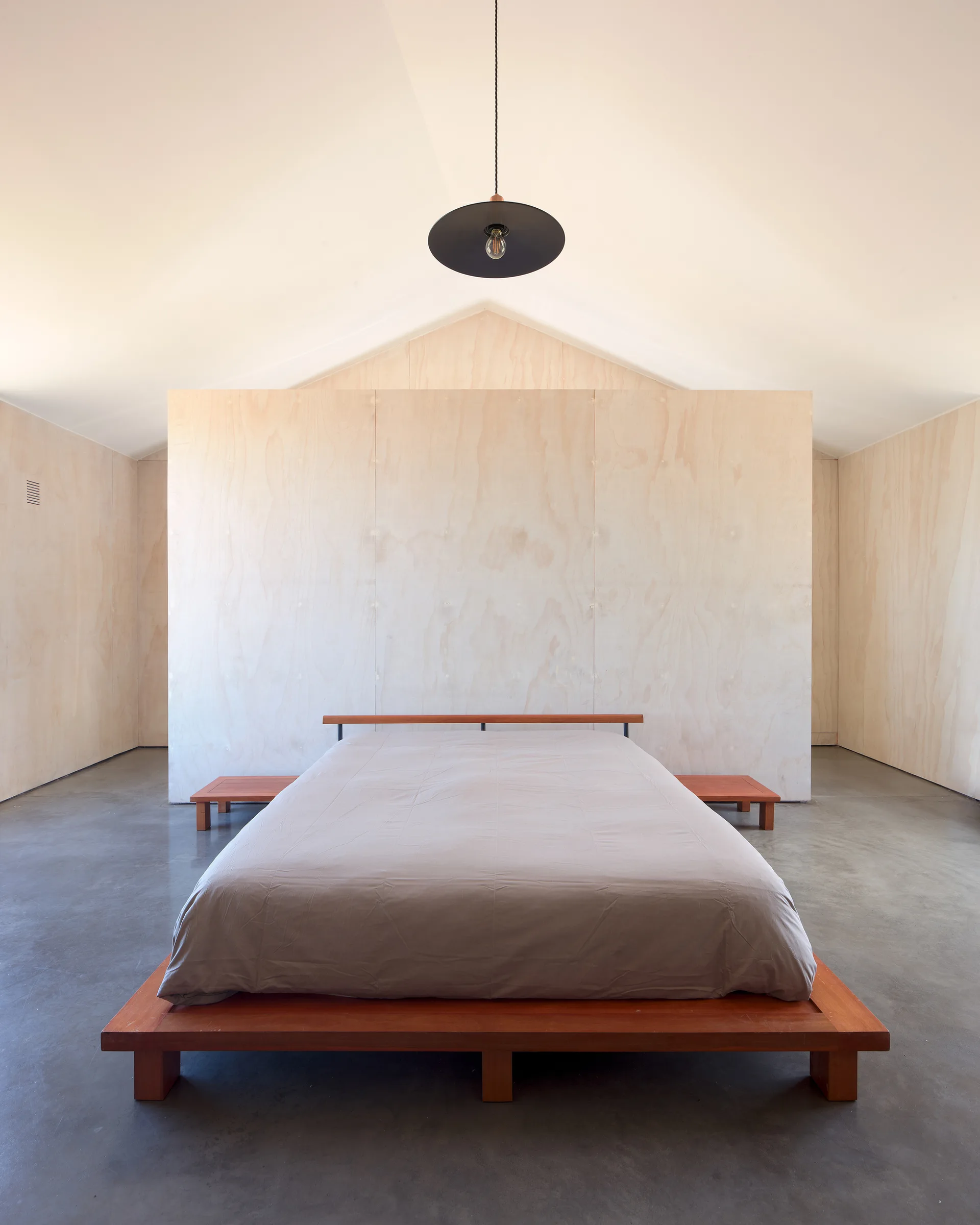 A minimalist bedroom features a wooden platform bed in front of a plywood partition wall with a concrete floor and a vaulted ceiling.