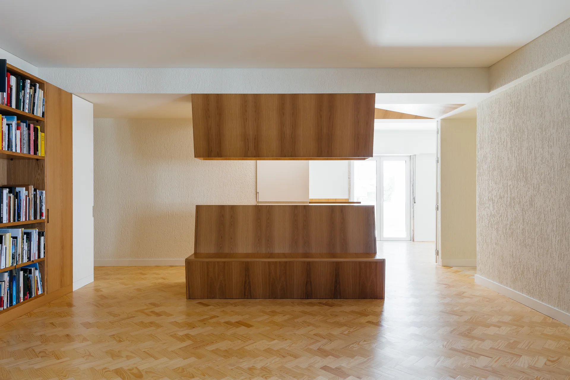 A minimalist room features a central floating wooden partition, built-in shelving, and light herringbone parquet floors against textured stucco walls.